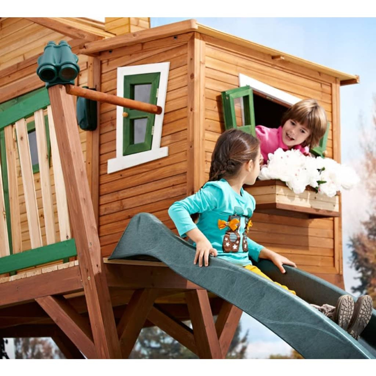 Zwei Kinder, eines auf einer Rutsche und eines in einem Fenster, auf einem Holzspielhaus.