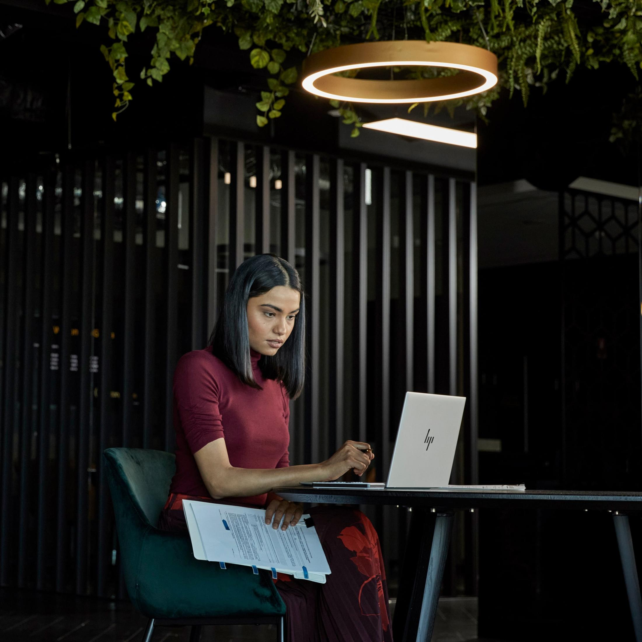 Kobieta w bordowym topie pracuje na laptopie. Jest w biurze z zielonym krzesłem.