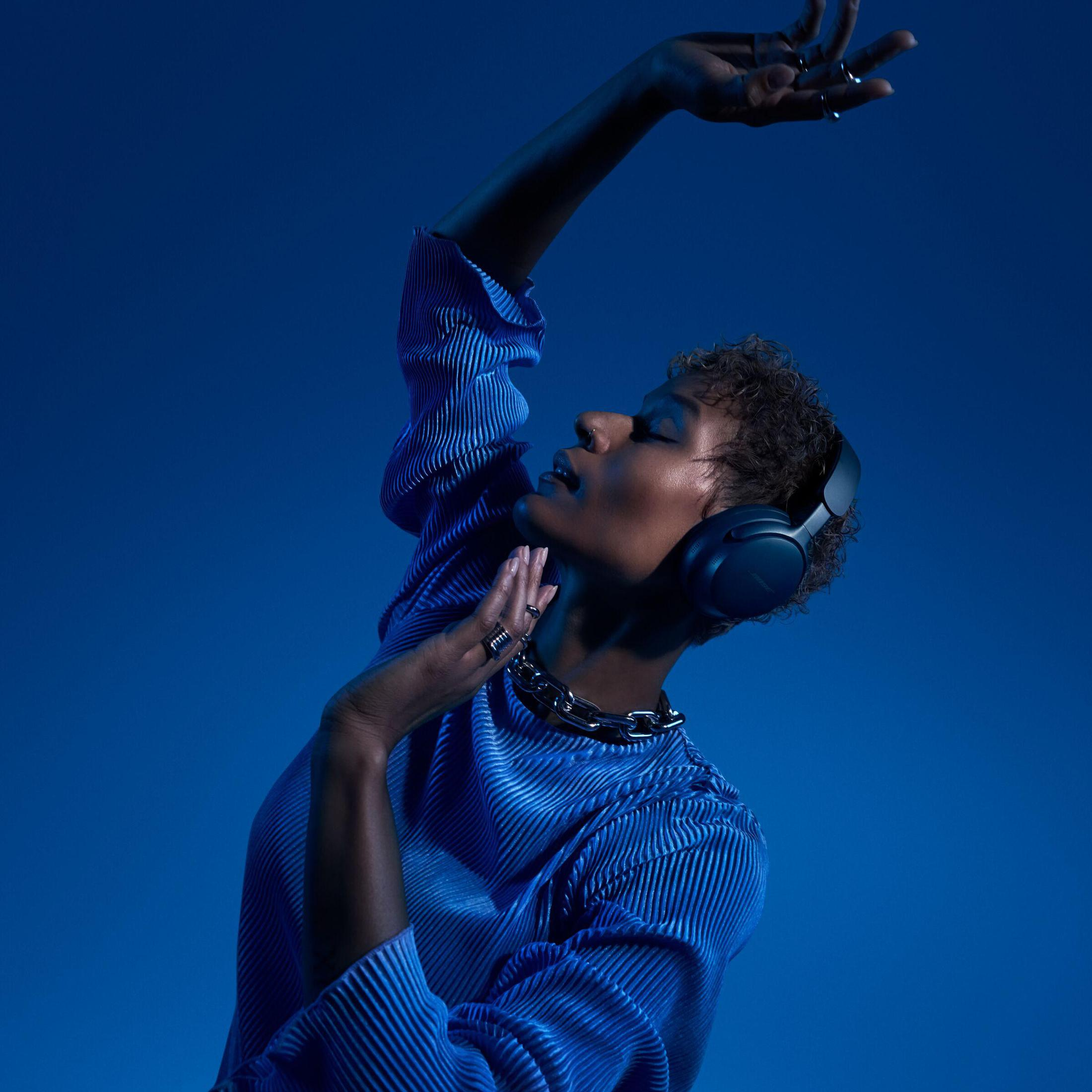 Kobieta ze słuchawkami pozuje z podniesionymi ramionami na niebieskim tle.