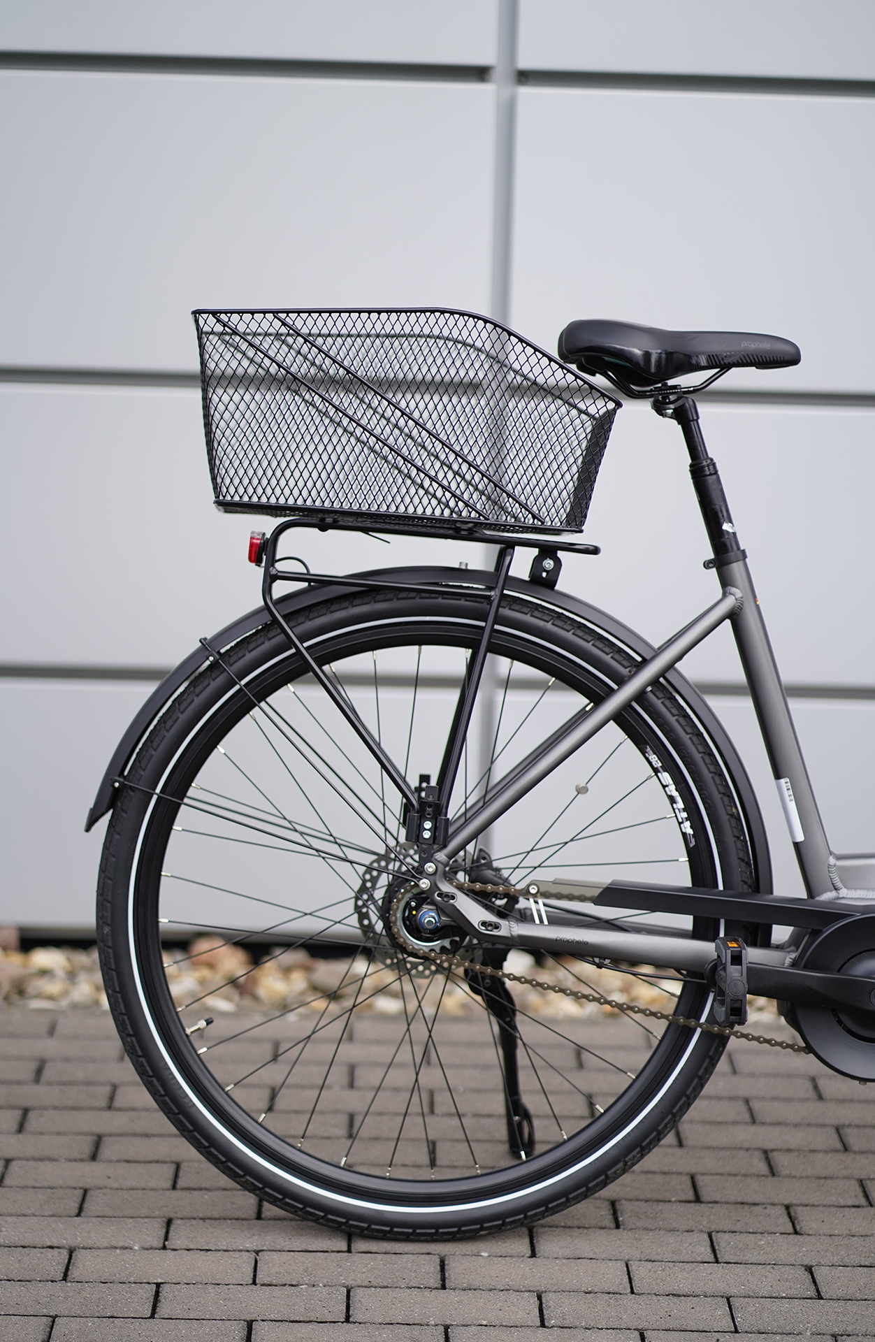 Graues Fahrrad mit schwarzem Korb. Steht auf Ziegelstein, graue Wand im Hintergrund.