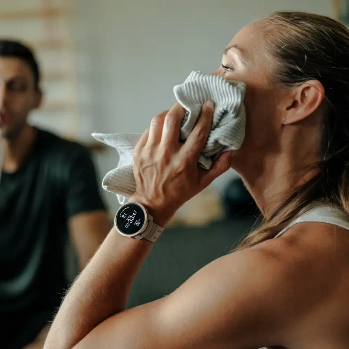 Kobieta w stroju do ćwiczeń wyciera twarz ręcznikiem, ma na ręce smartwatch.