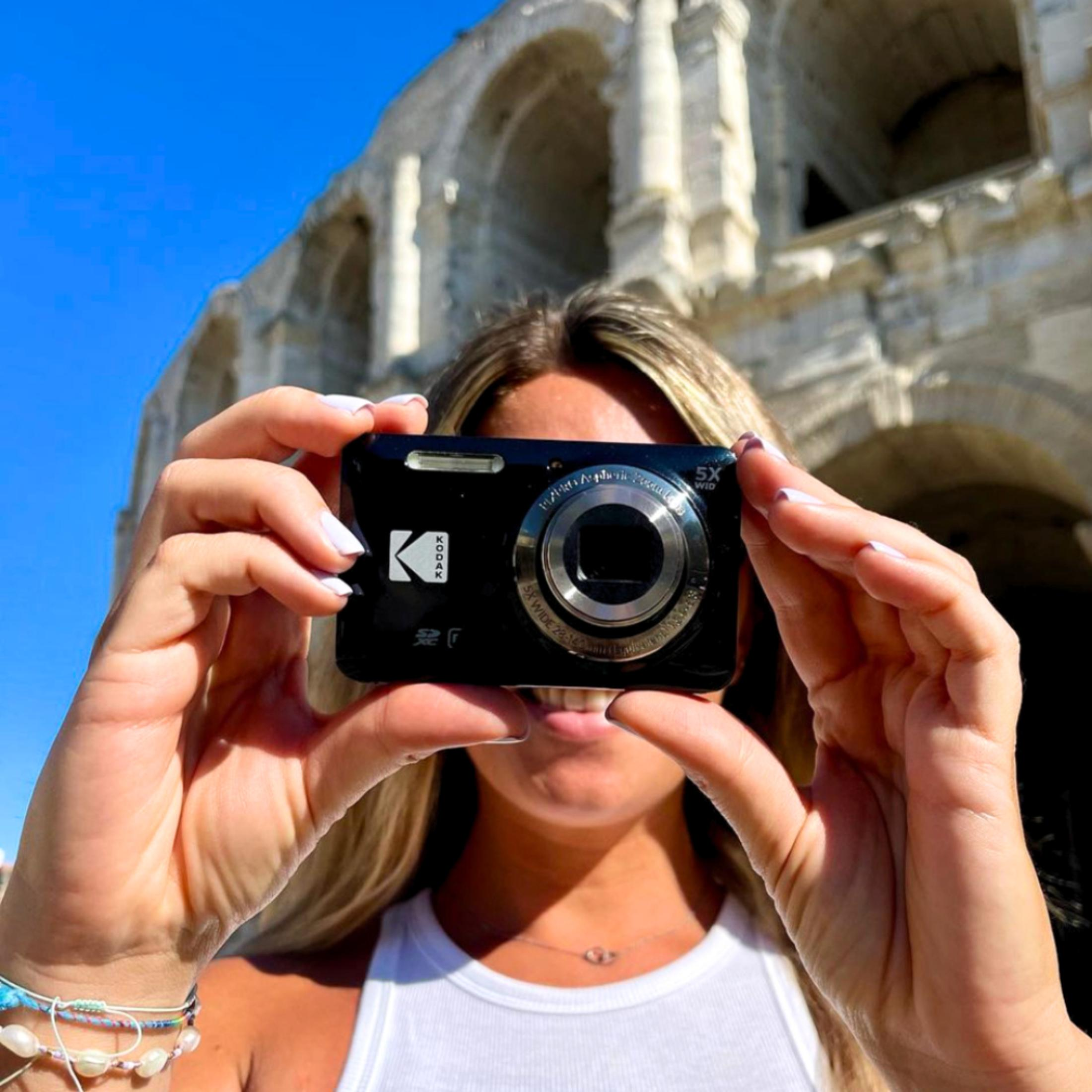 Kobieta trzymająca czarny aparat Kodak przed twarzą, w tle starożytny budynek.