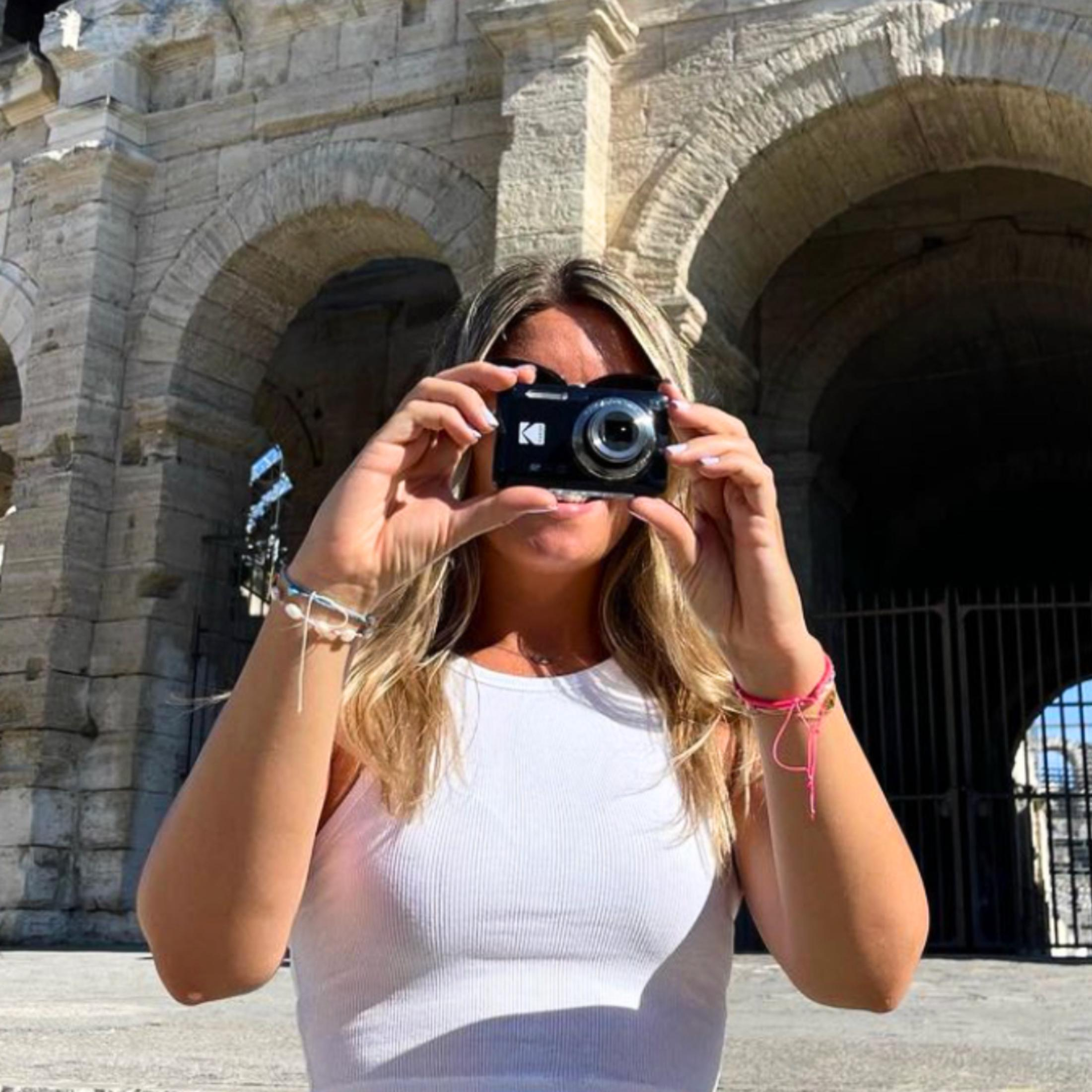 Kobieta trzyma aparat Kodak przed twarzą, za nią starożytny budynek.