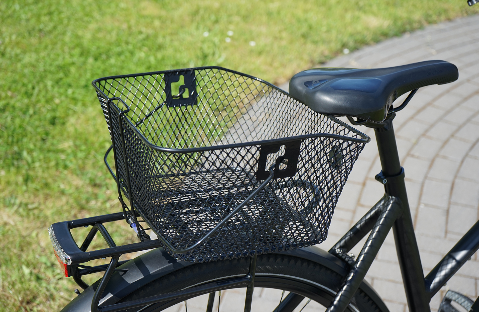 Schwarzes Fahrrad mit Drahtkorb am Heck, grünes Gras und Pflasterung.