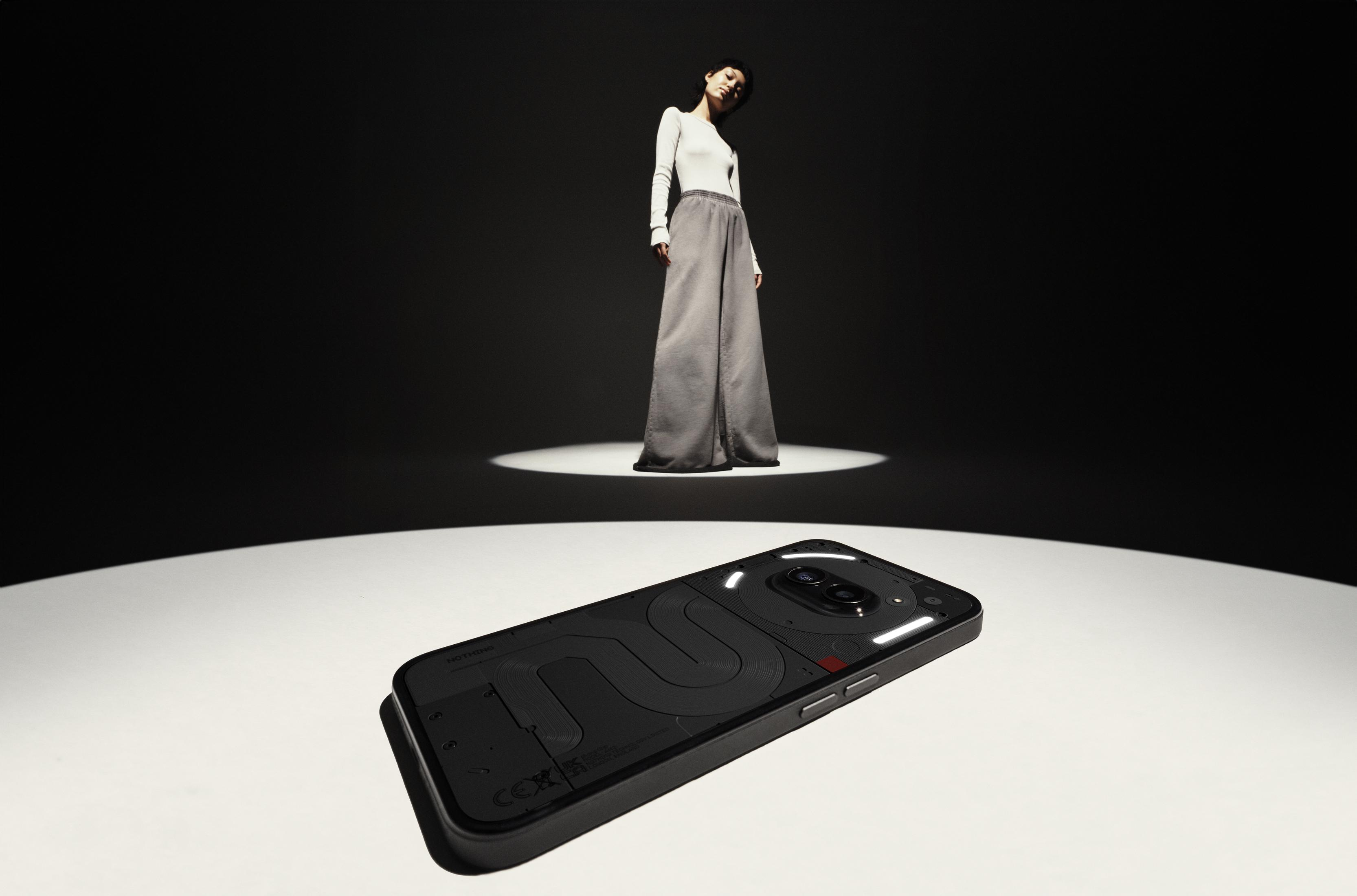 Eine Frau in einem weißen Oberteil und grauer Hose steht über einem schwarzen Telefon auf einer weißen Fläche.