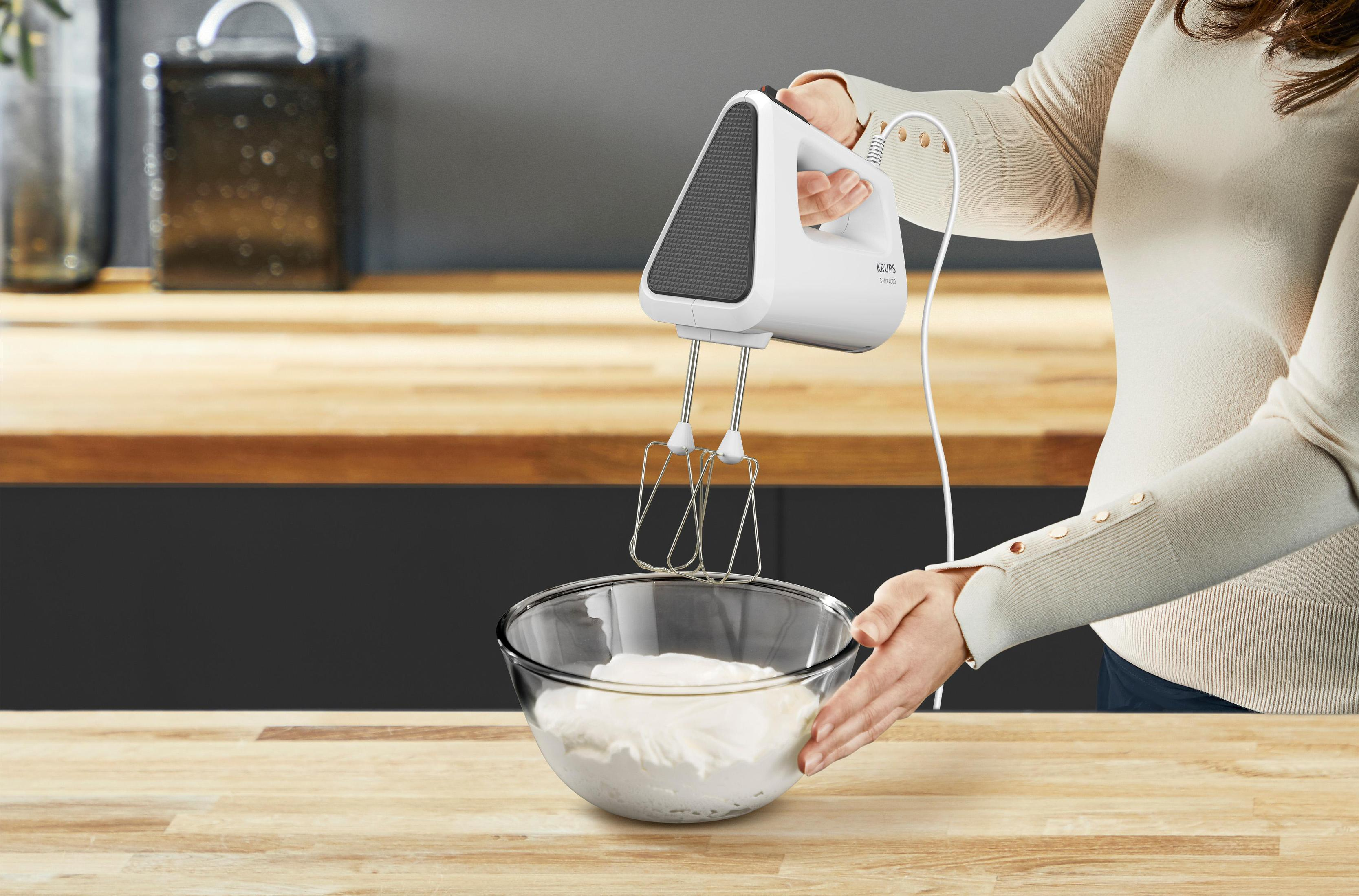 Een vrouw die een witte handmixer gebruikt boven een kom slagroom op een houten aanrecht.