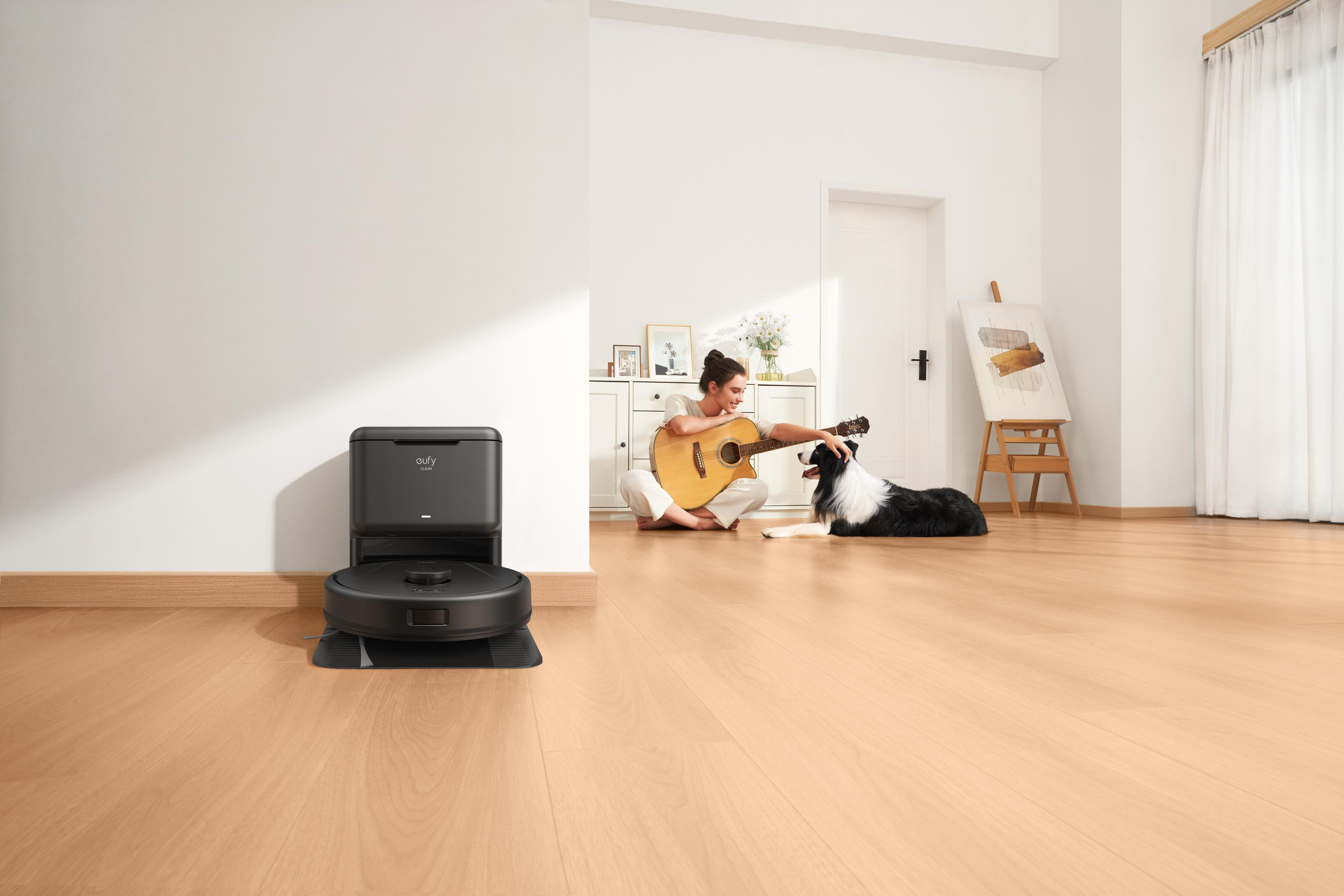 Kobieta gra na gitarze z psem obok robota sprzątającego. Wnętrze z białymi ścianami.