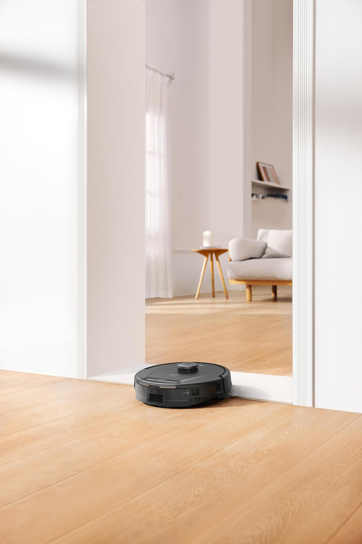 Robot sprzątający na drewnianej podłodze, widziany przez drzwi. Pomieszczenie w tle.