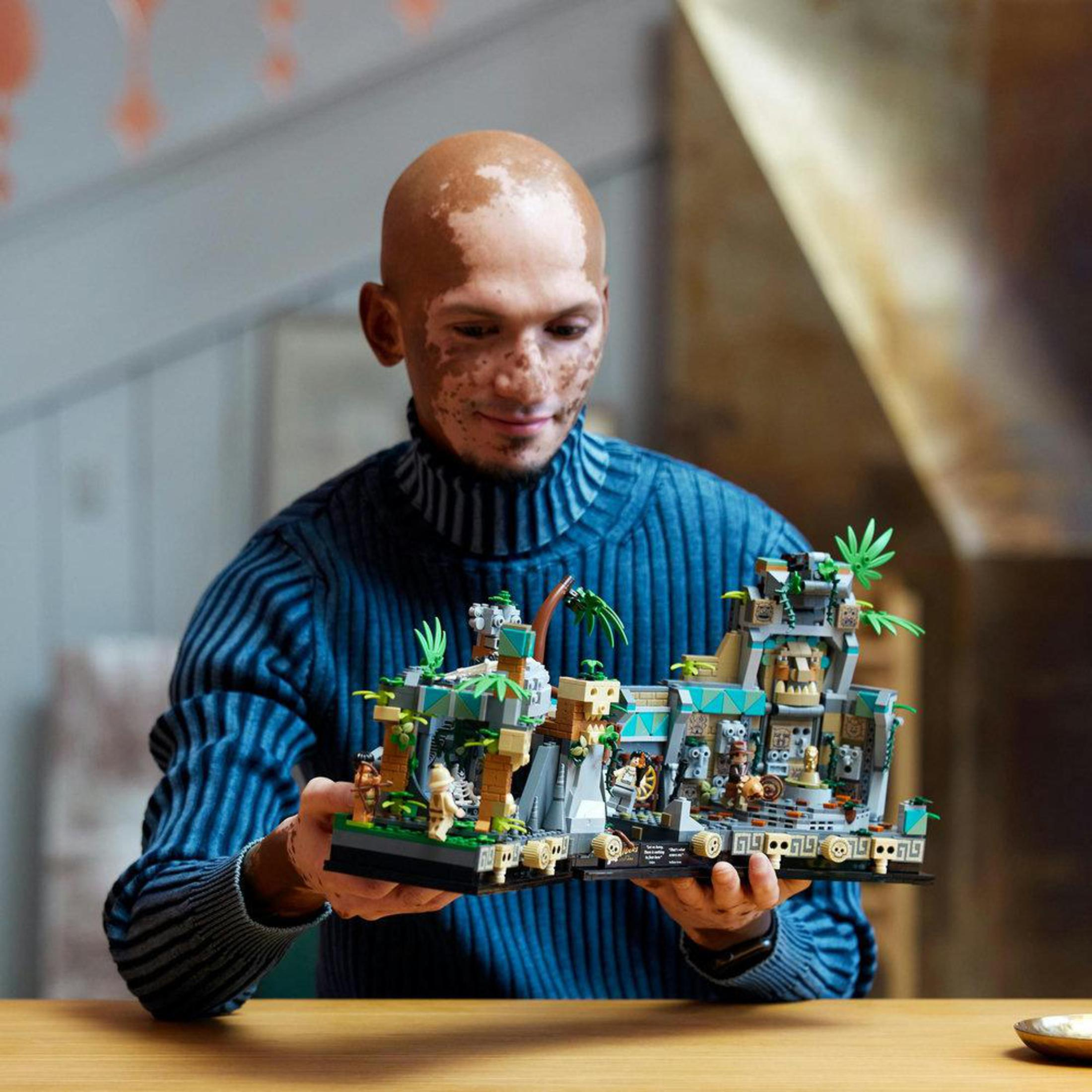 Ein Mann hält ein Lego-Set eines Tempels mit Figuren. Er lächelt, hat helle Haut und Haarausfall.