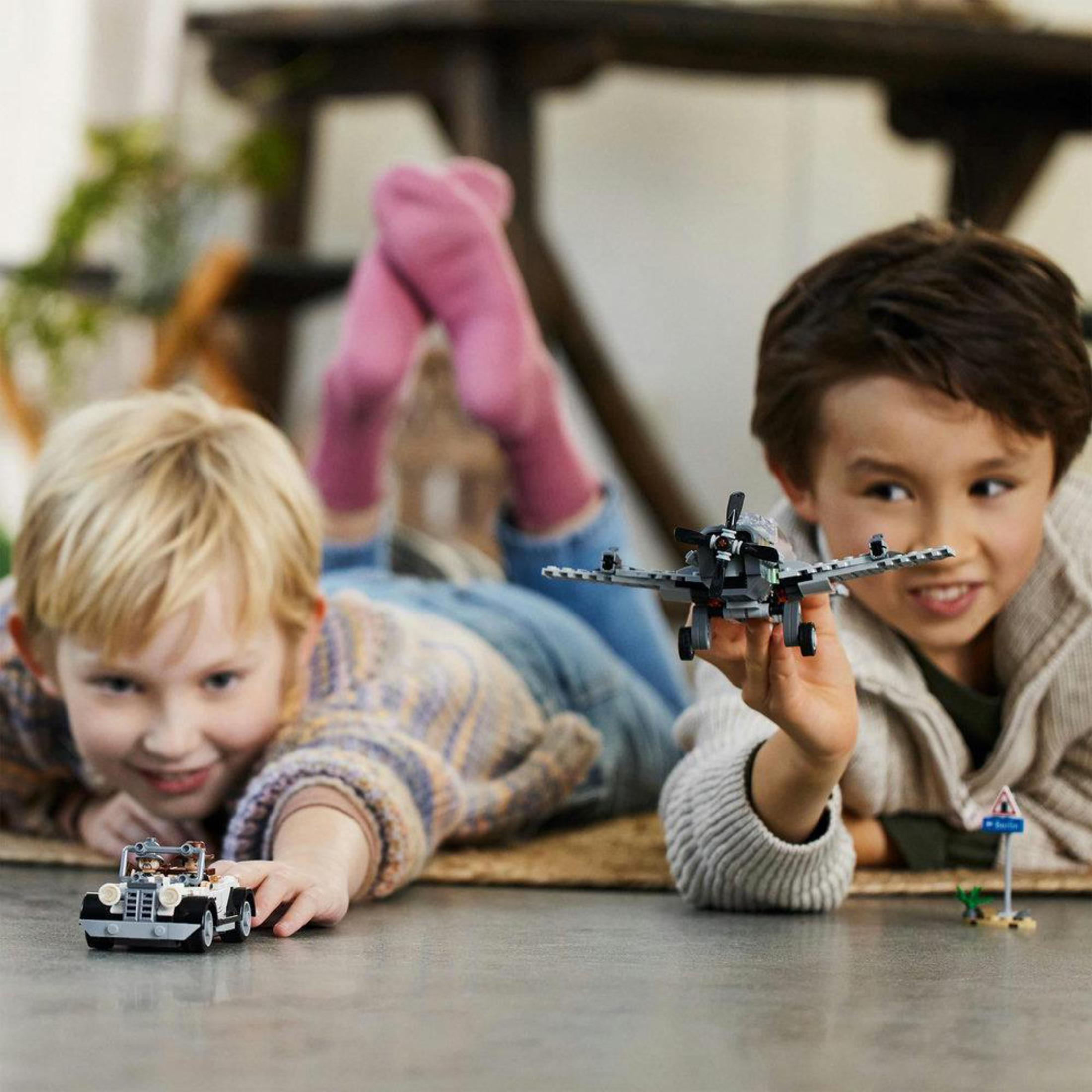 Dos niños jugando con coches de juguete y un avión. Están tumbados en una alfombra, sonriendo.