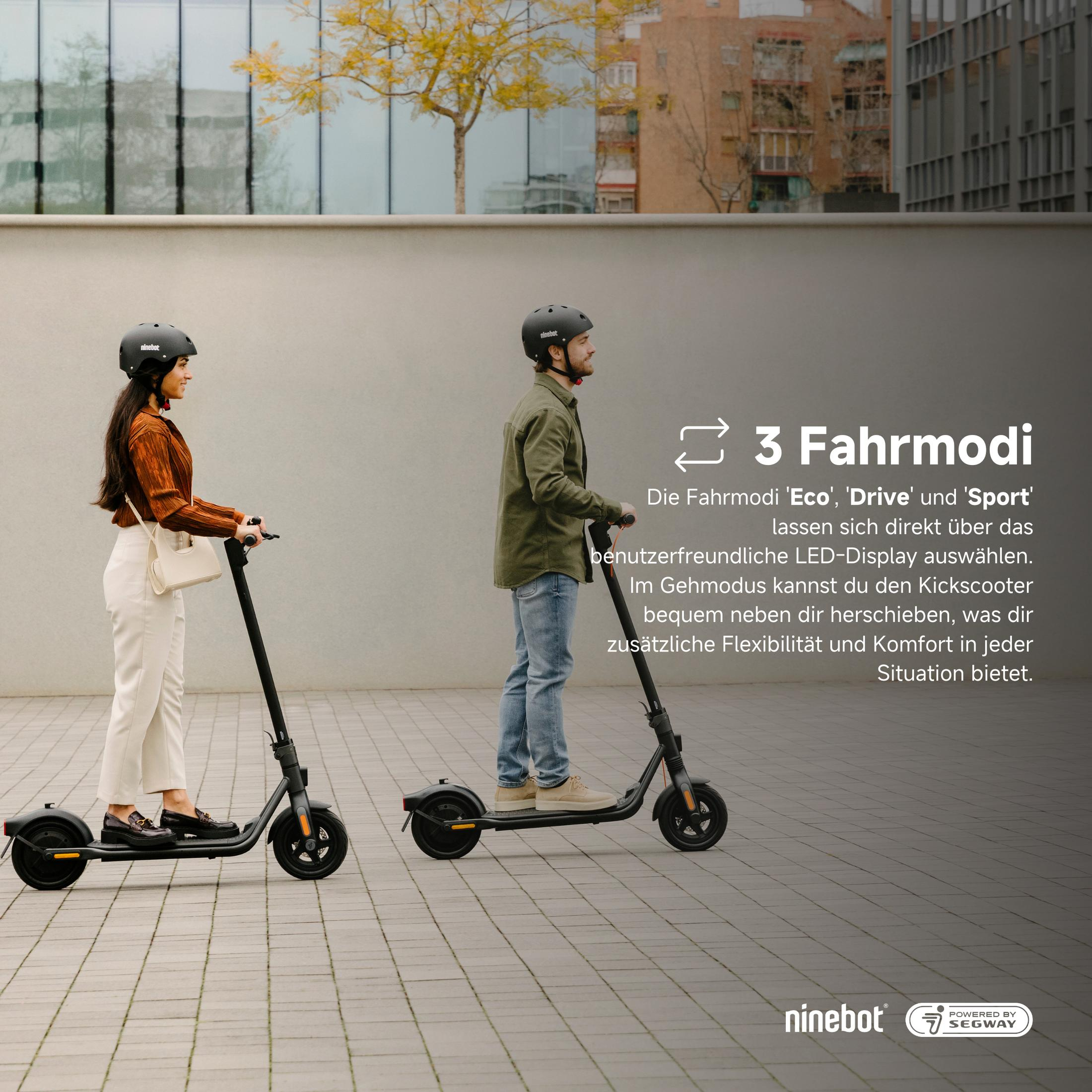 Zwei Personen mit Helmen fahren mit schwarzen Ninebot-Elektrorollern auf einem gepflasterten Bereich mit einem Gebäude und einem Baum im Hintergrund.