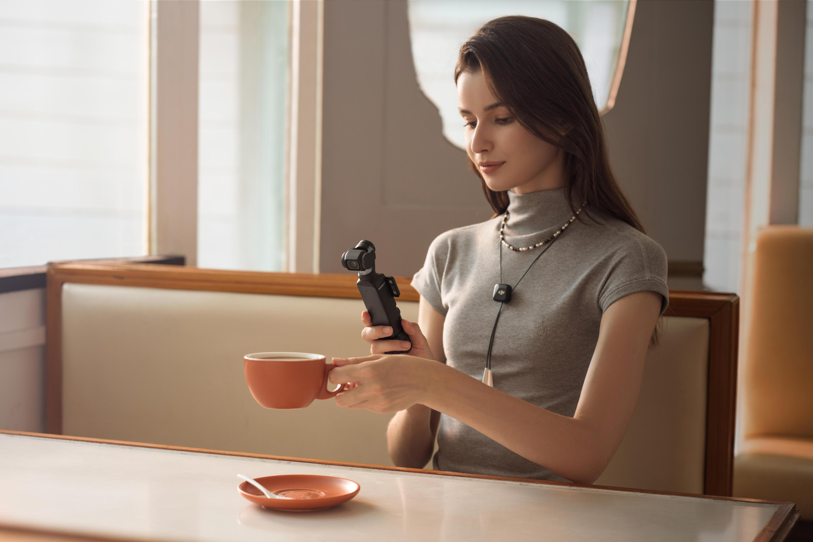 Kobieta trzymająca ciemne urządzenie, filiżankę i spodek. Siedzi w pomieszczeniu, światło z okien.