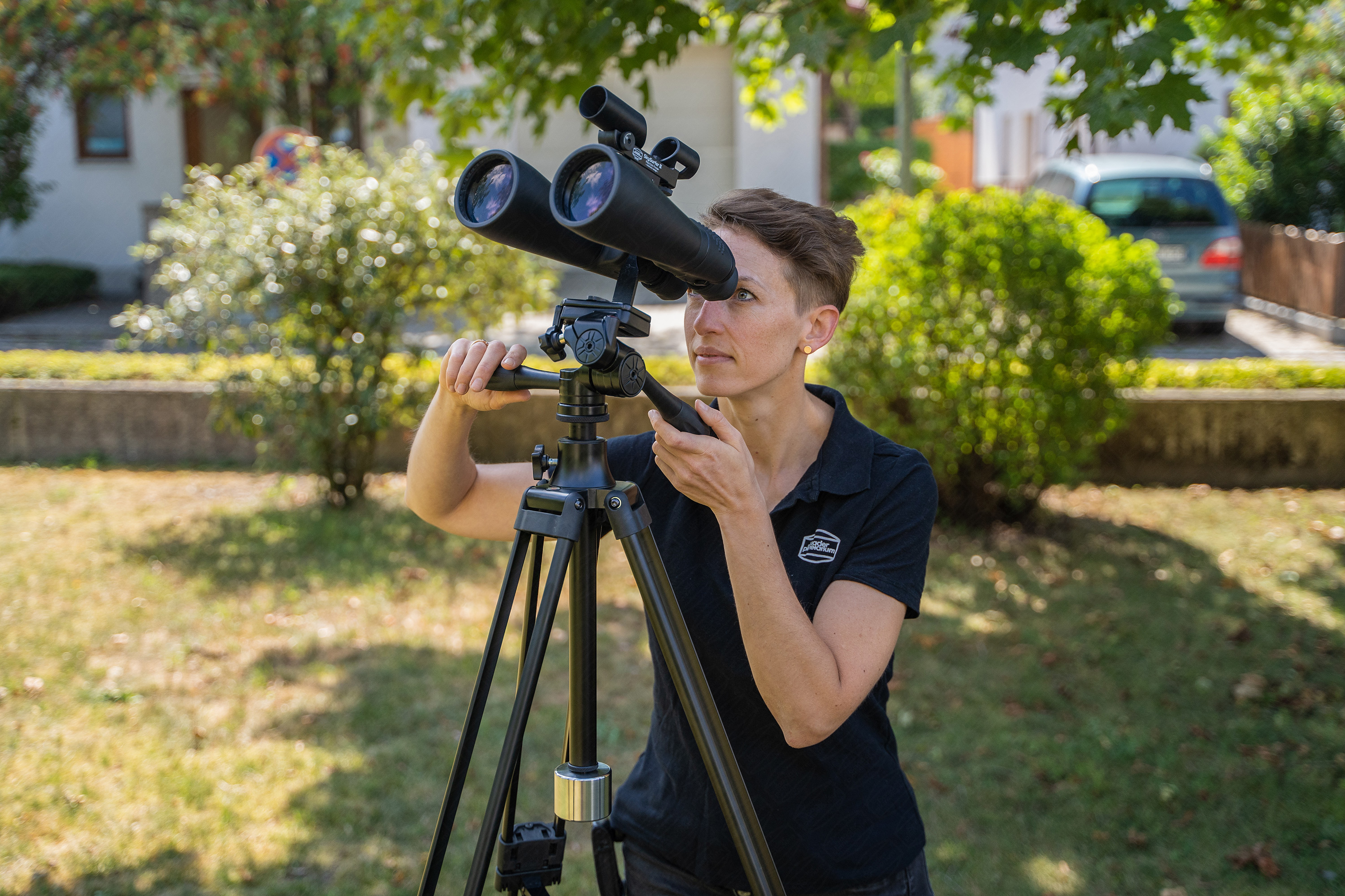 Frau benutzt Fernglas auf einem Stativ im Freien. Grünes Gras und Bäume im Hintergrund.