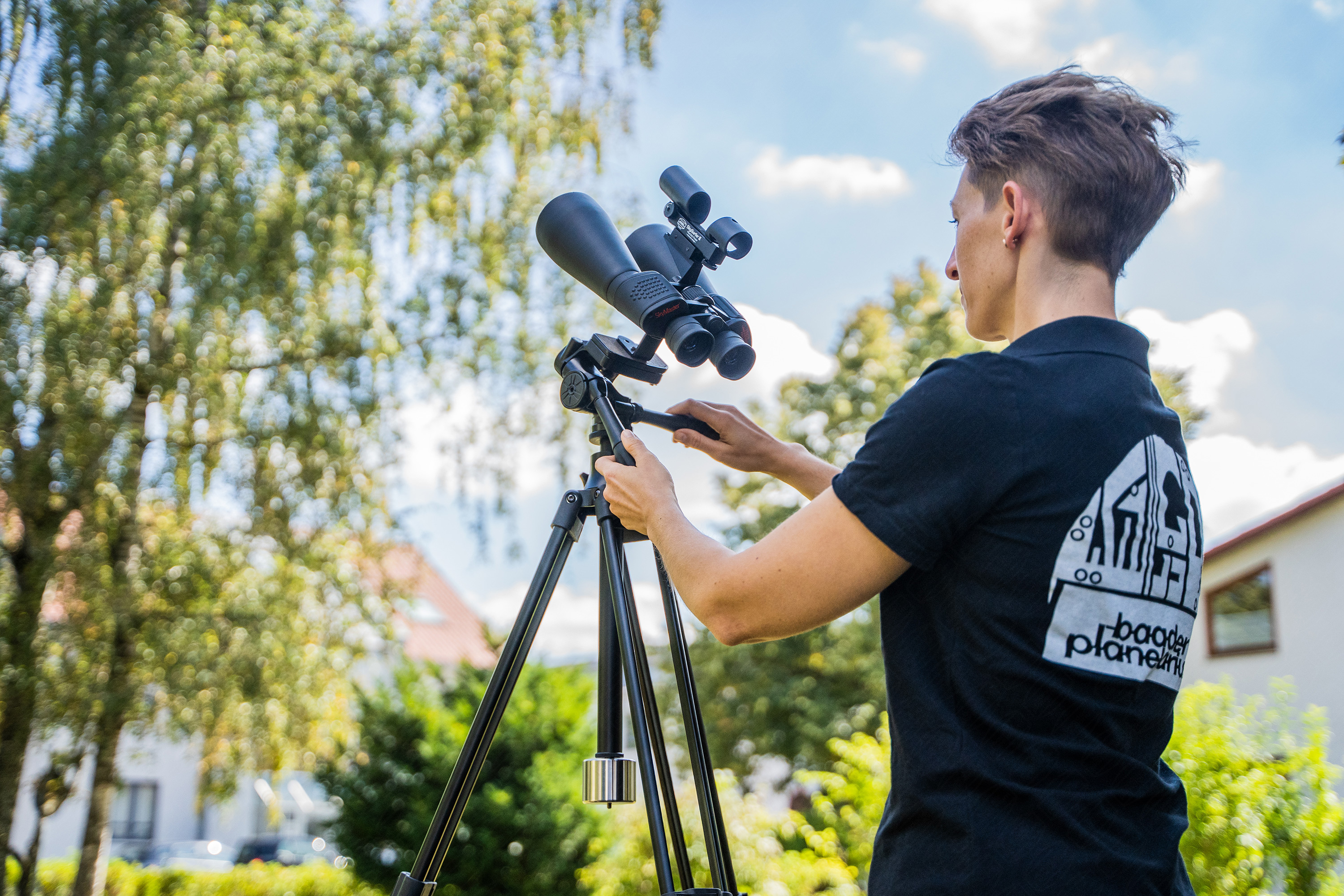 Person justiert Fernglas auf Stativ im Freien mit Bäumen im Hintergrund.