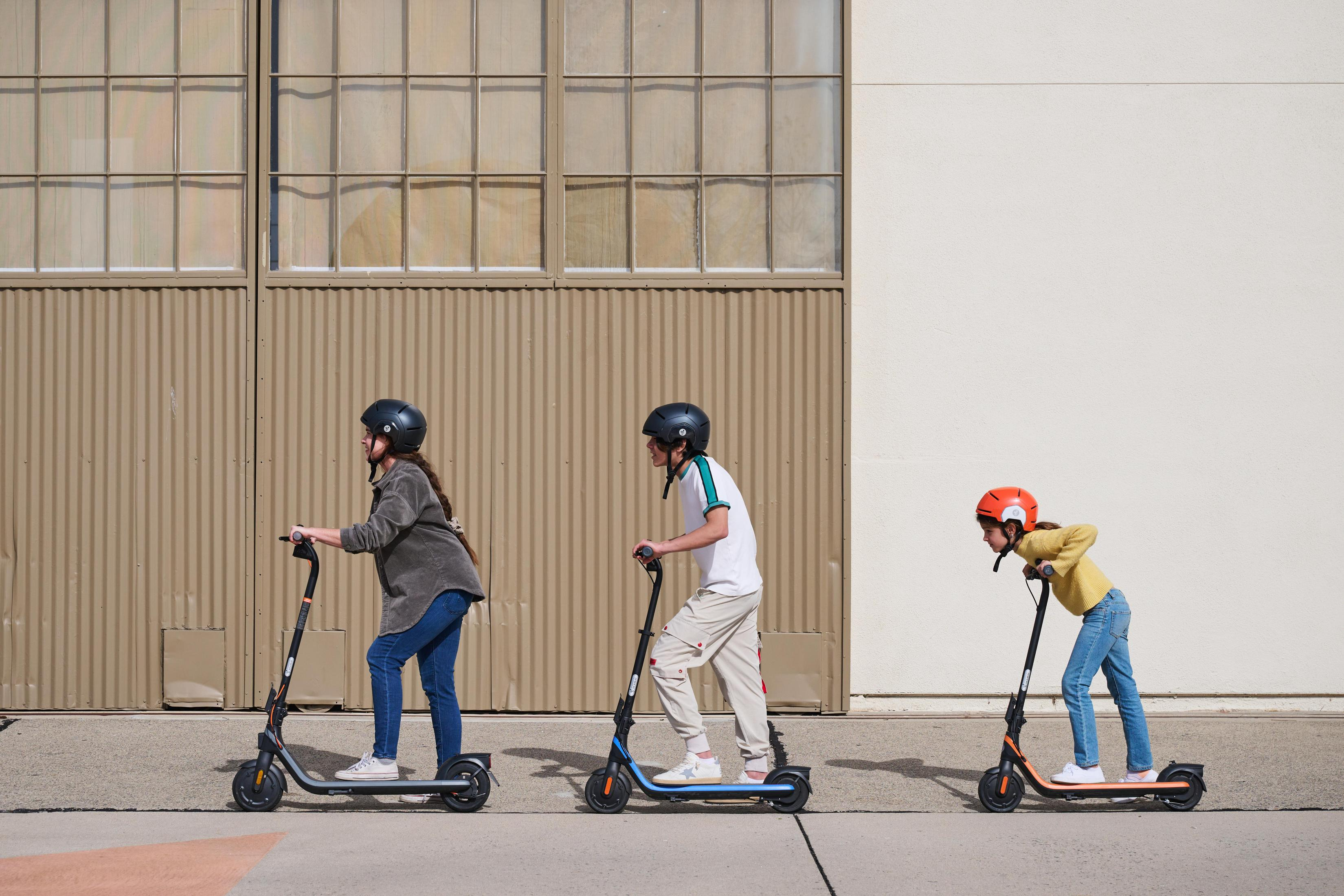 Troje dzieci jeździ na hulajnogach w słoneczny dzień. Mają na sobie kaski i stoją w rzędzie.