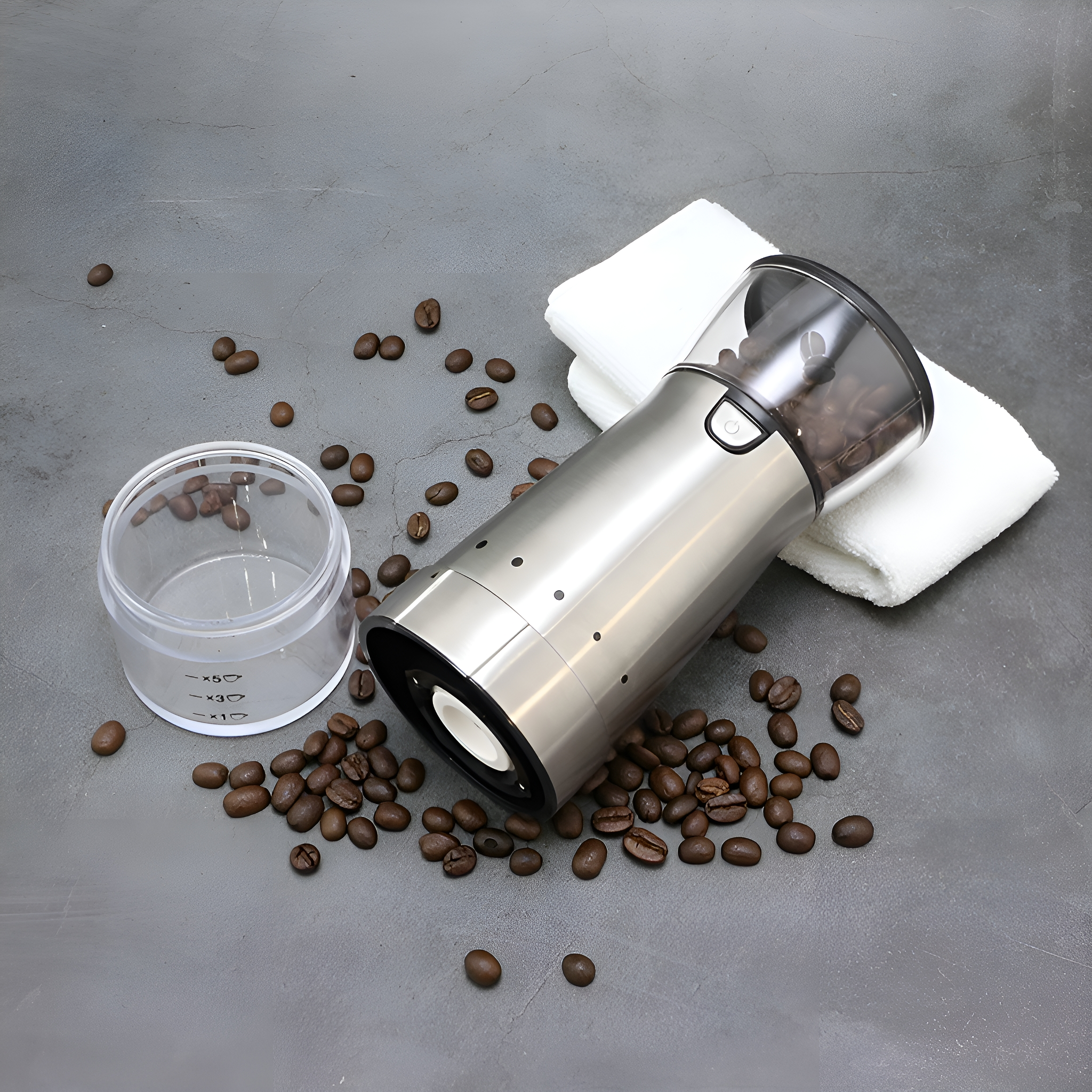 Edelstahl-Kaffeemühle mit Bohnen, Messbecher und weißem Tuch auf grauer Oberfläche.
