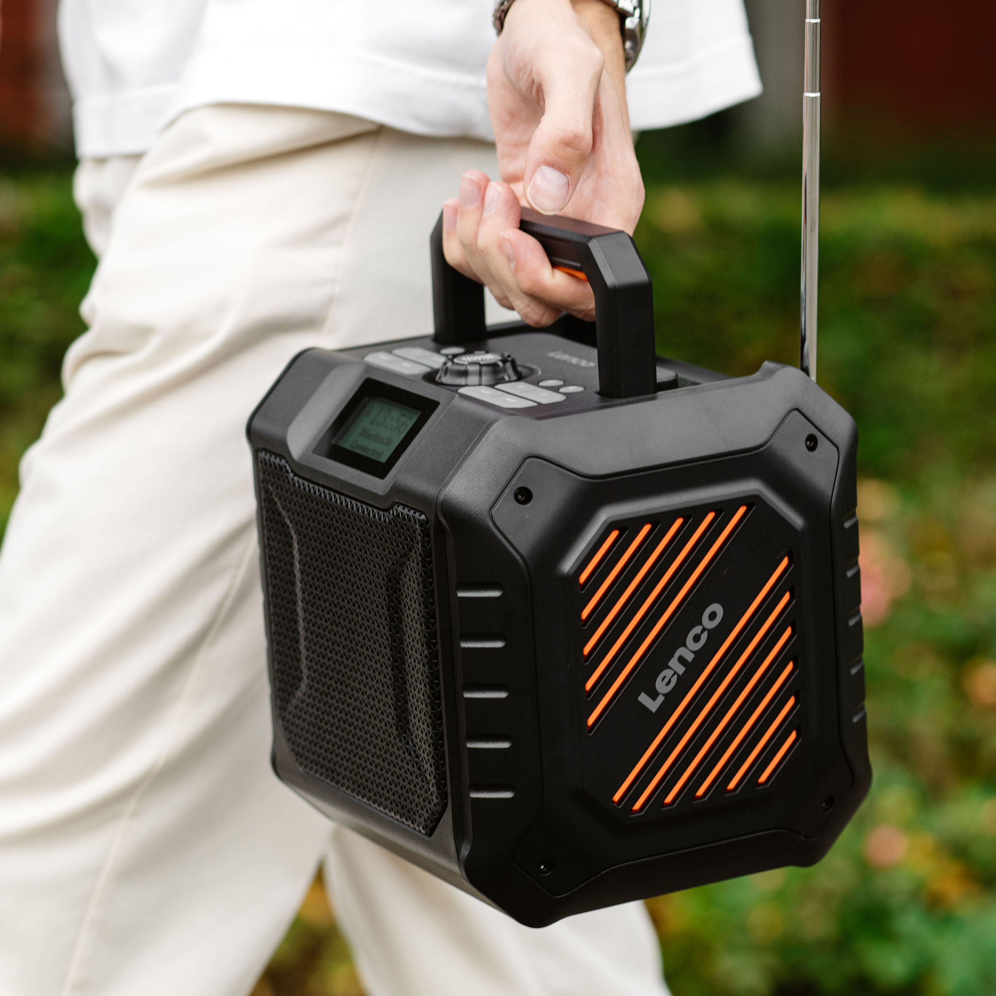Osoba trzymająca czarno-pomarańczowe przenośne radio Lenco. Tło to rozmyte zielone liście.