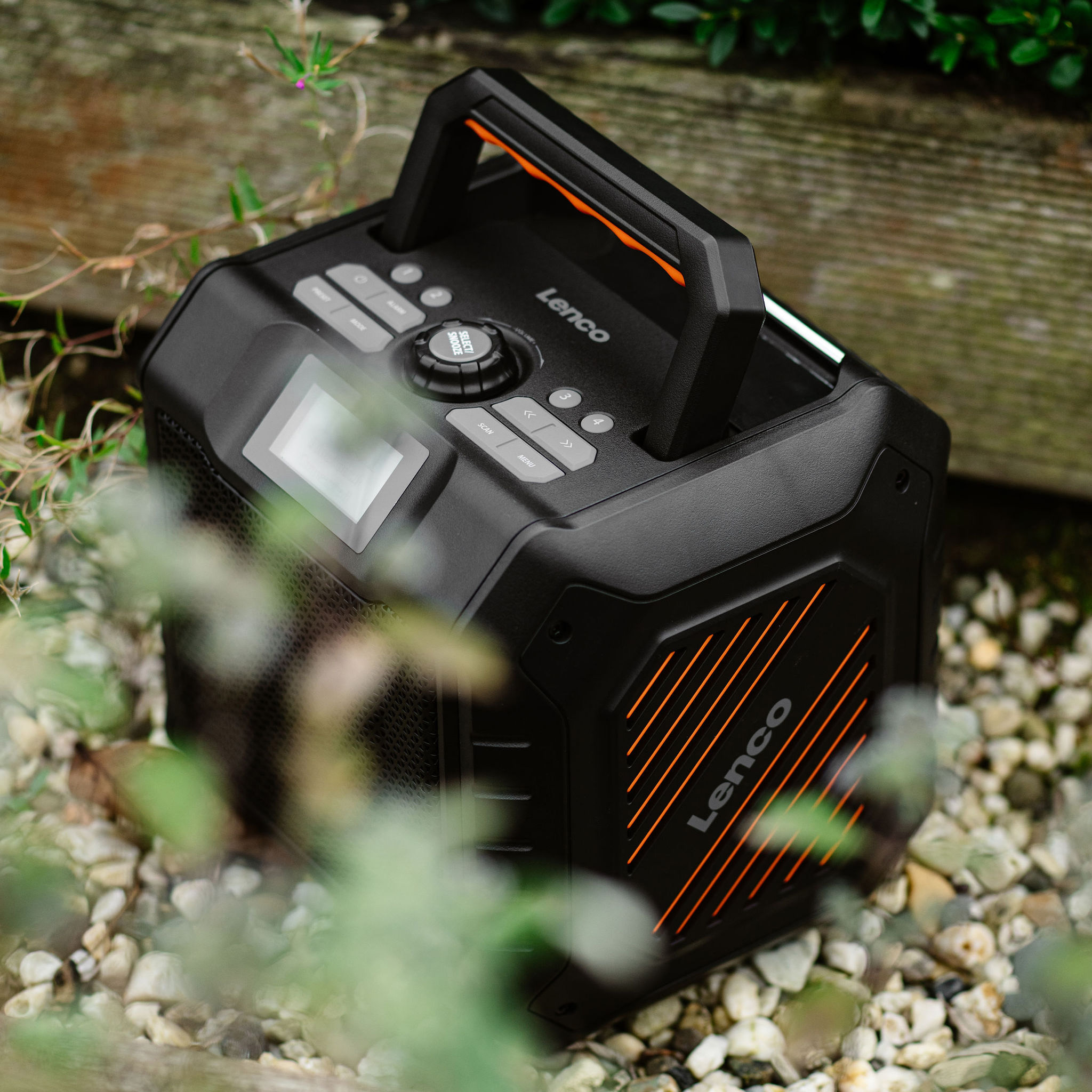 Czarny głośnik Lenco z pomarańczowymi paskami. Otoczony zielenią, na kamieniach.