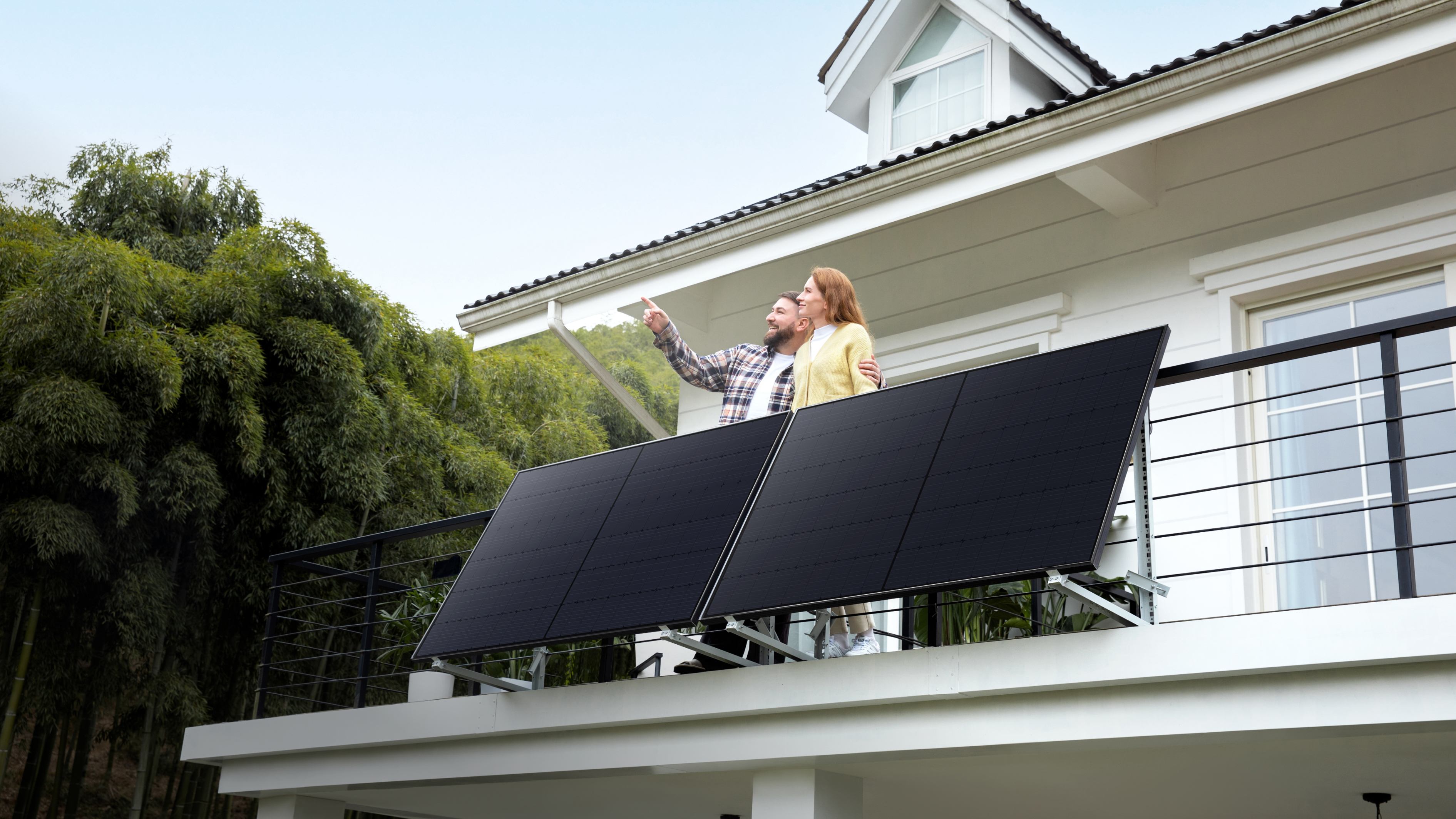 Una pareja en un balcón, señalando paneles solares. Árboles verdes y una casa forman el fondo.