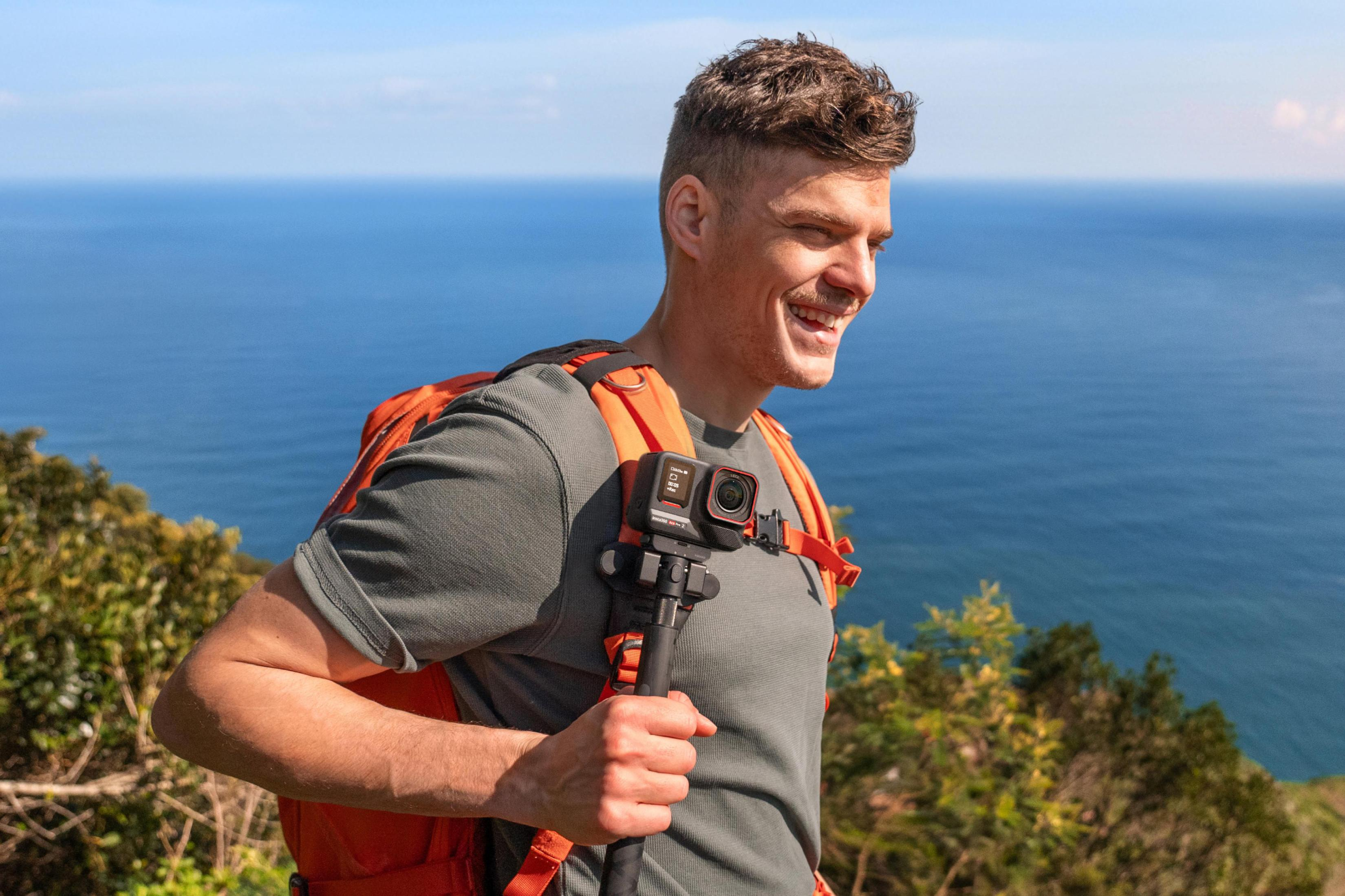 Mężczyzna z plecakiem filmuje kamerą na górze z błękitnym morzem.
