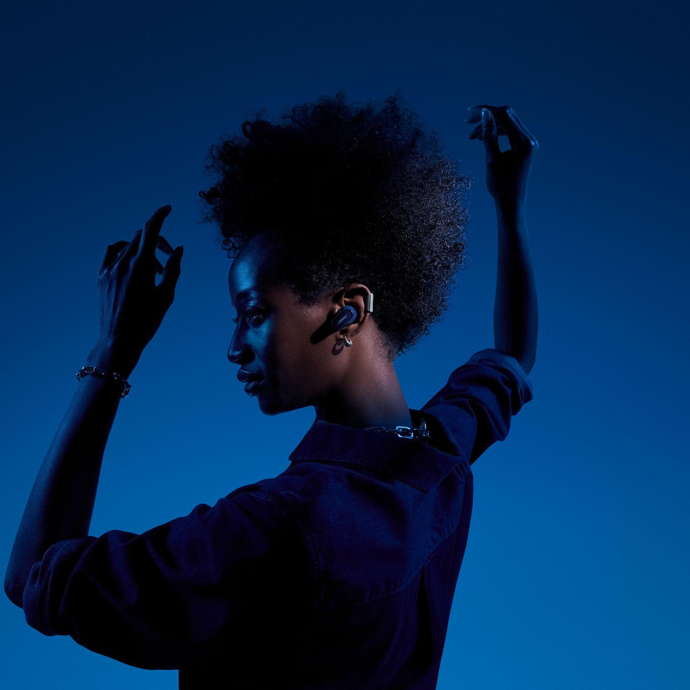 Kobieta z afro nosząca słuchawki, podniesione ręce, niebieskie tło. Widok z boku, cienie. Słuchawki są granatowe.