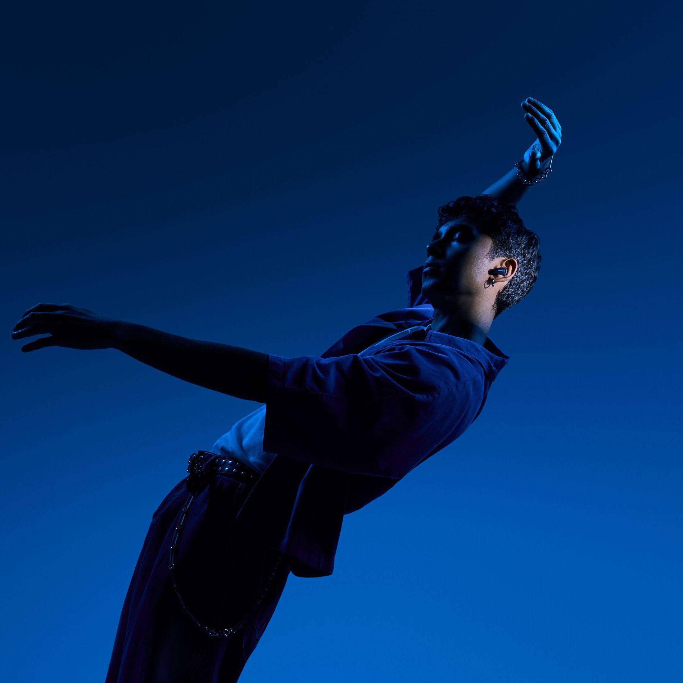 Osoba w niebieskim stroju tańczy z podniesionymi ramionami, ze słuchawkami, na niebieskim tle.