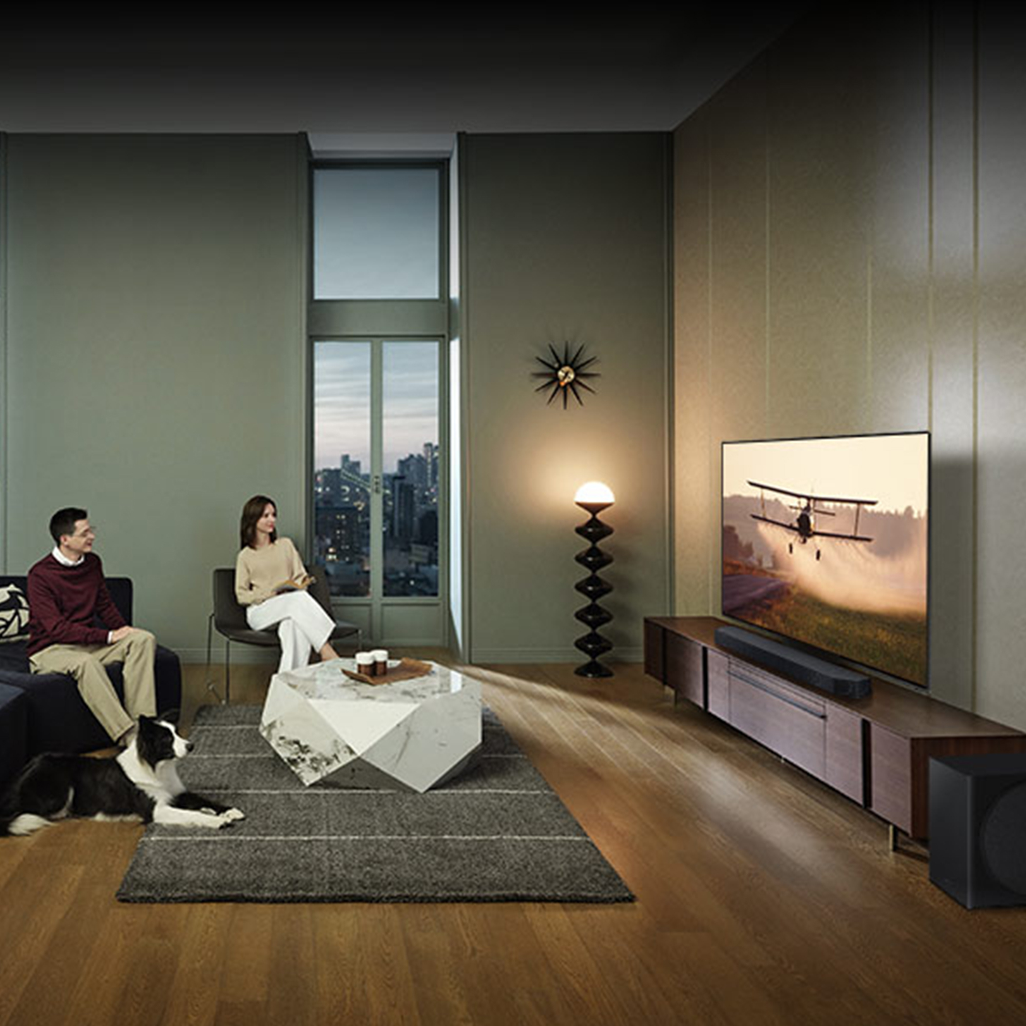 Ein Wohnzimmer mit Fernseher. Ein Paar sitzt auf einem Sofa. Ein Hund liegt auf dem Boden.