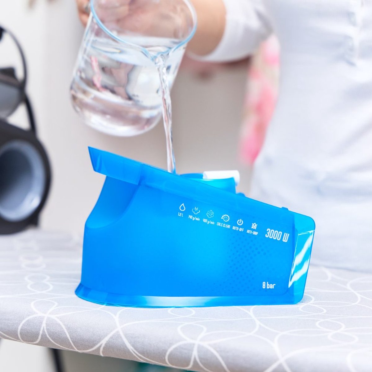 Una persona versa acqua in una base di ferro blu su un'asse da stiro, con testo e icone.