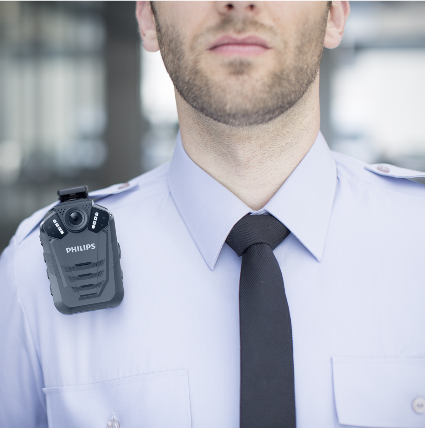 Ein Mann in Uniform mit schwarzer Krawatte und Körperkamera. Die Kamera trägt die Aufschrift 'PHILIPS'. Der Hintergrund ist unscharf.