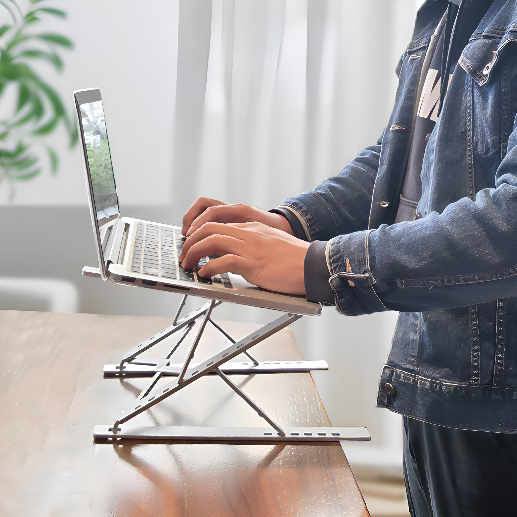 Person verwendet Laptop auf silbernem Laptopständer auf Holztisch. Jeansjacke sichtbar.