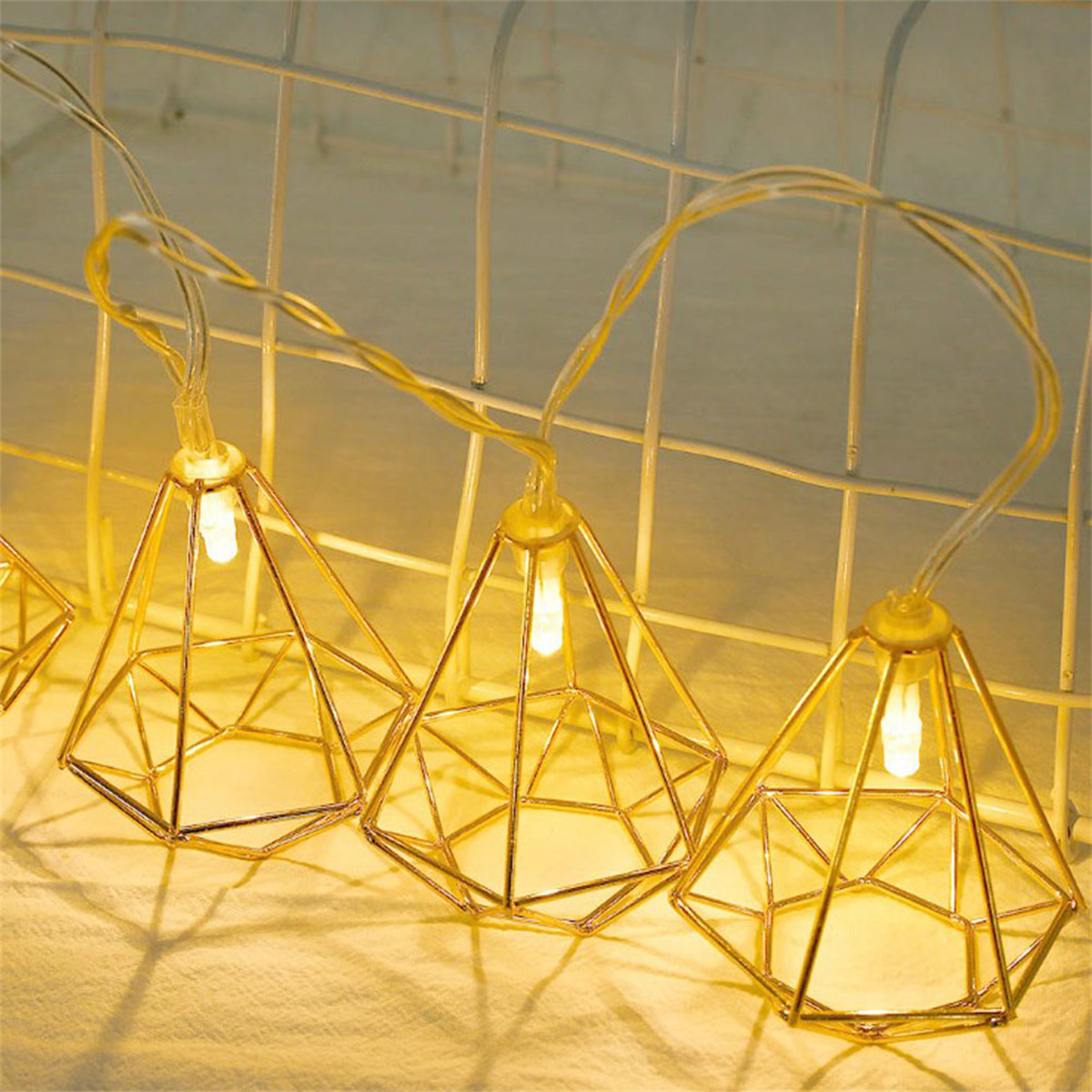 Goldene geometrische Leuchten, beleuchtet und hängend vor weißem Metallgitterhintergrund.