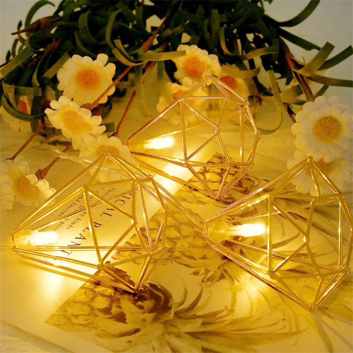 Goldene geometrische Dekorationslichter mit Blumen im Hintergrund. Die Lichter befinden sich auf einer hellen Oberfläche.