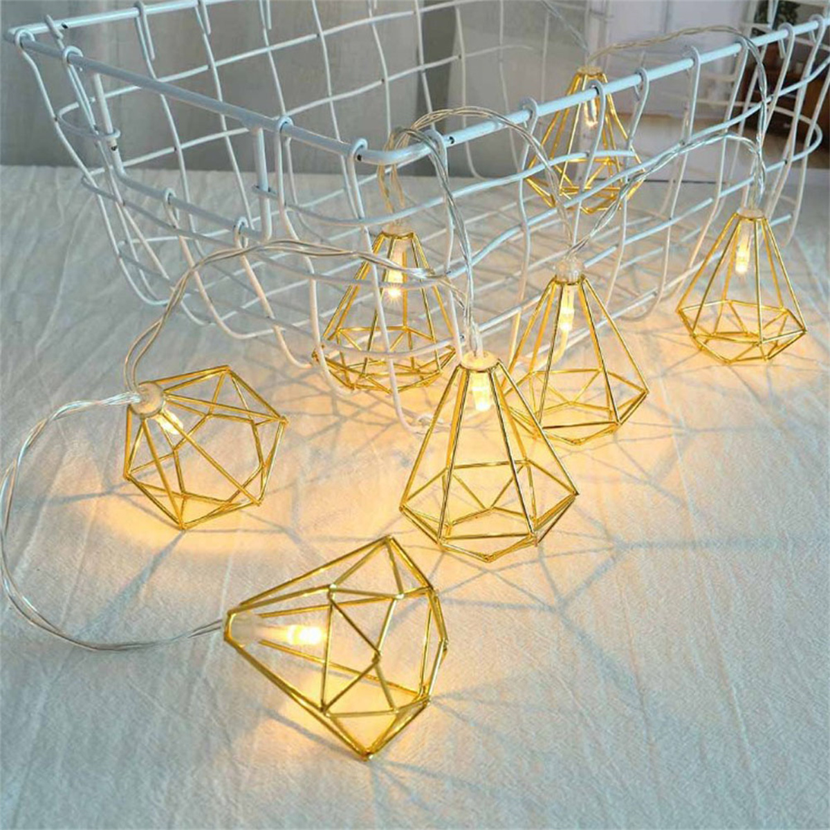 Goldfarbene geometrische Lichter hängen an einem Draht in einem weißen Korb, auf weißer Oberfläche.