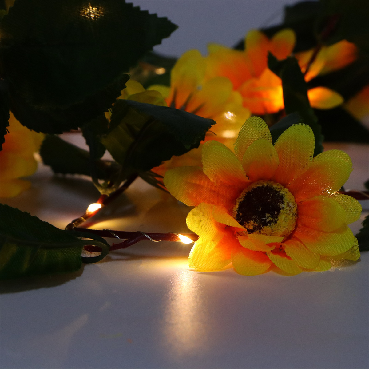 Nahaufnahme von künstlichen Sonnenblumen mit Lichtern auf weißer Oberfläche. Grüne Blätter sichtbar.