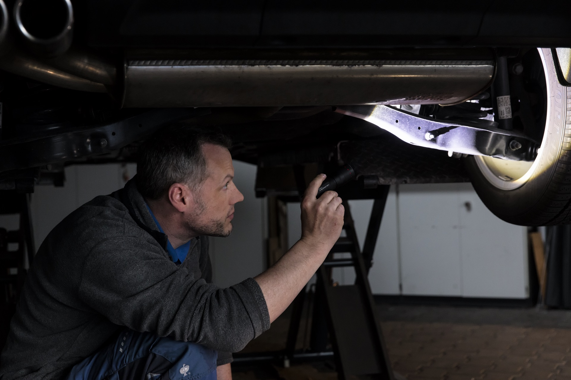 Mężczyzna w szarym swetrze sprawdza spód samochodu z latarką w garażu.