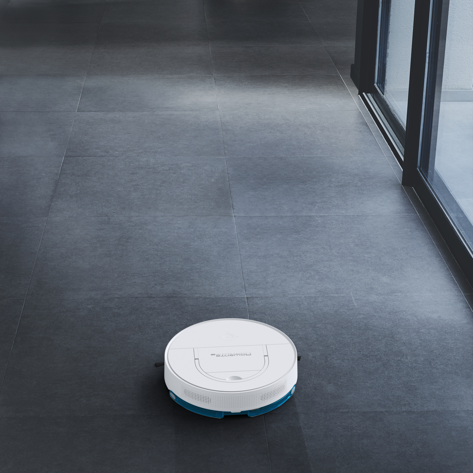 Aspirapolvere robotico su pavimento piastrellato grigio. Finestre visibili sullo sfondo. Interni puliti e moderni.