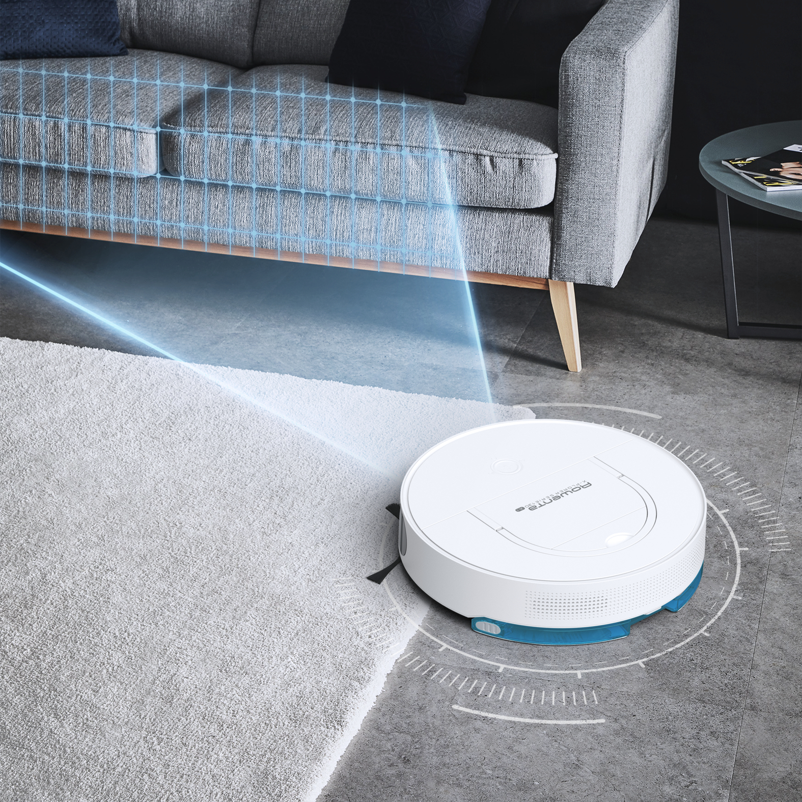 Un aspirapolvere robot bianco su pavimento grigio, vicino a un divano grigio e un tappeto bianco.
