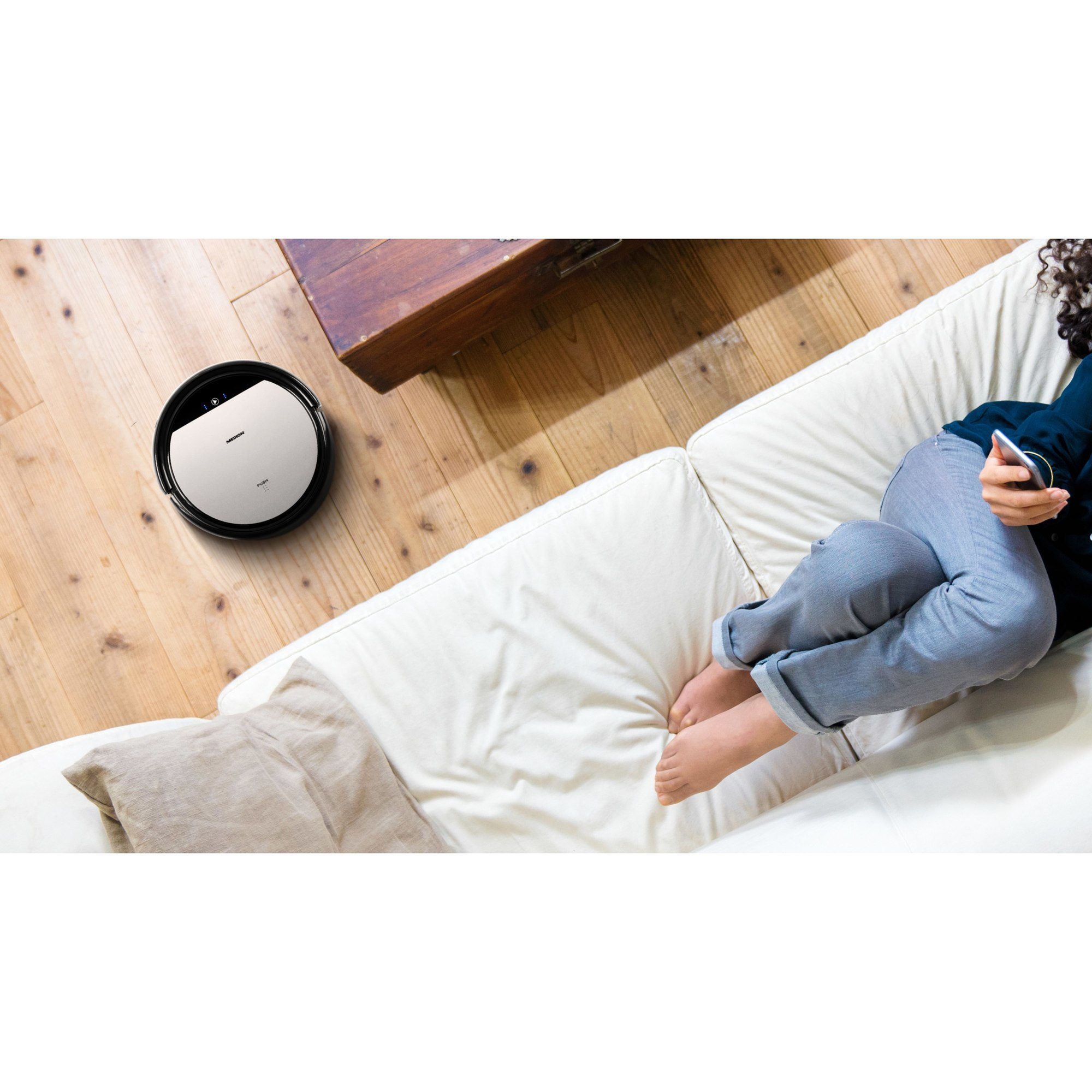 Roboter-Staubsauger auf Holzboden, neben weißem Sofa. Person benutzt Telefon.