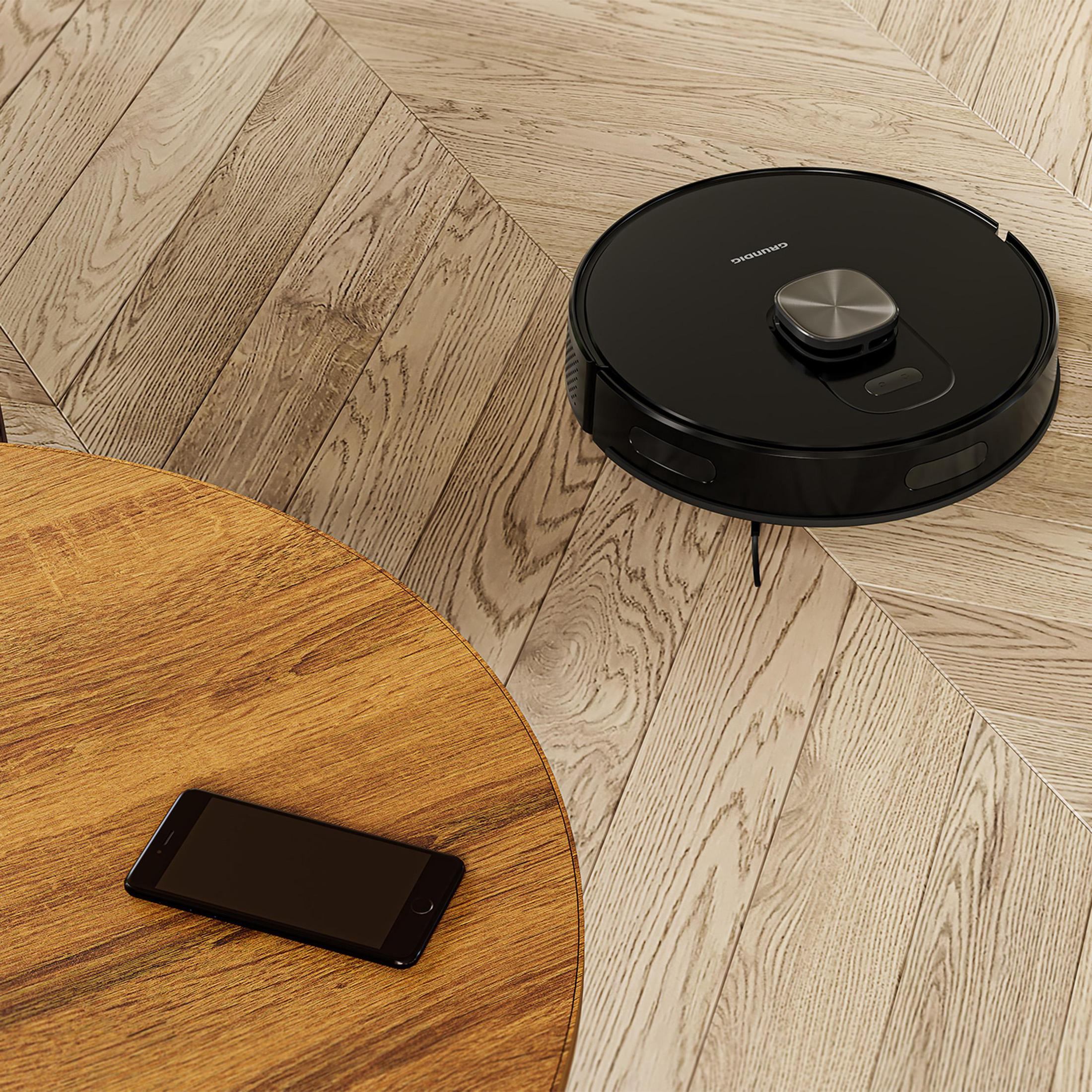 Schwarzer Roboter-Staubsauger auf Holzboden. Es gibt zwei Holztische und ein Telefon.