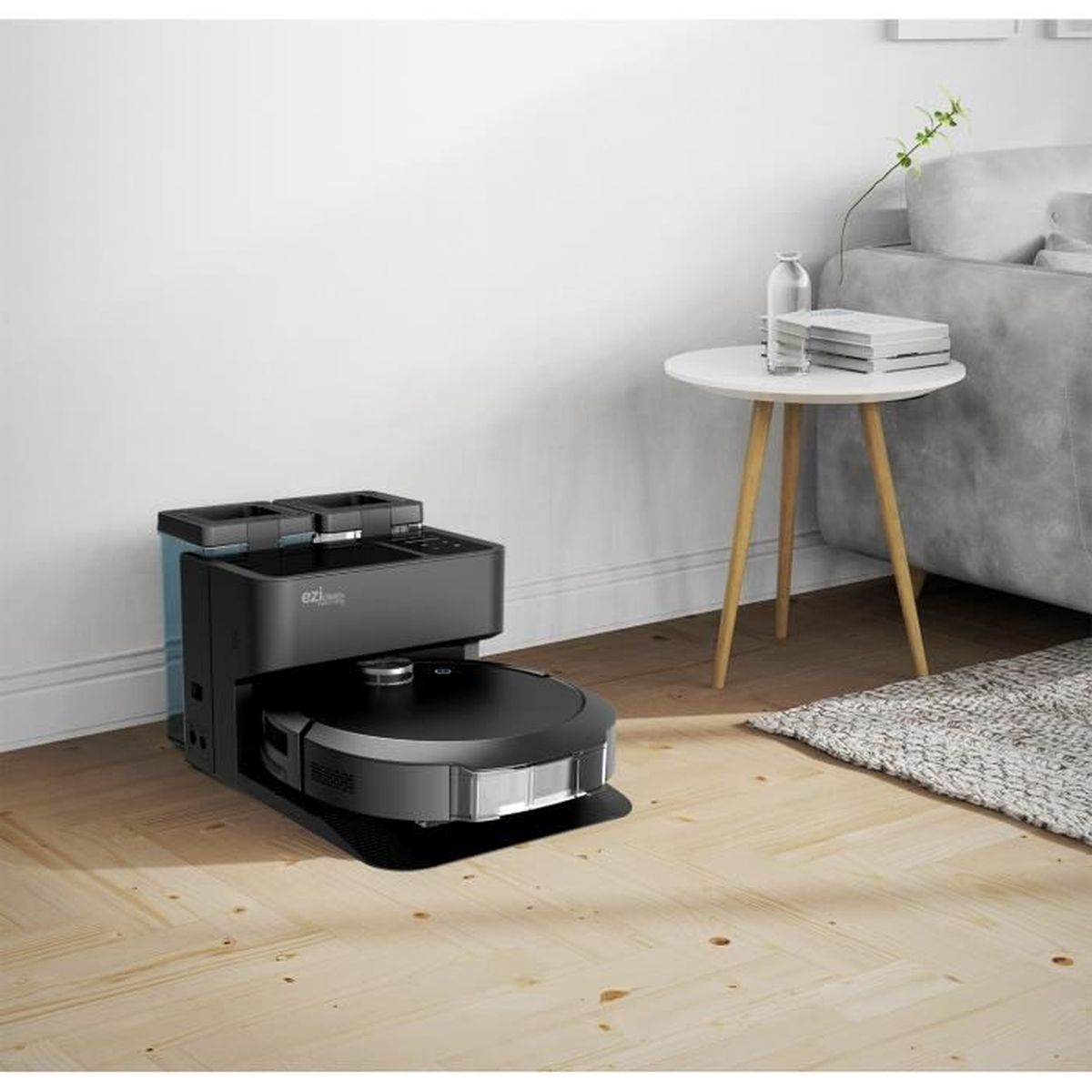 Robot odkurzający w pokoju. Jest czarny, ze stacją ładującą. Widoczna drewniana podłoga i białe ściany.