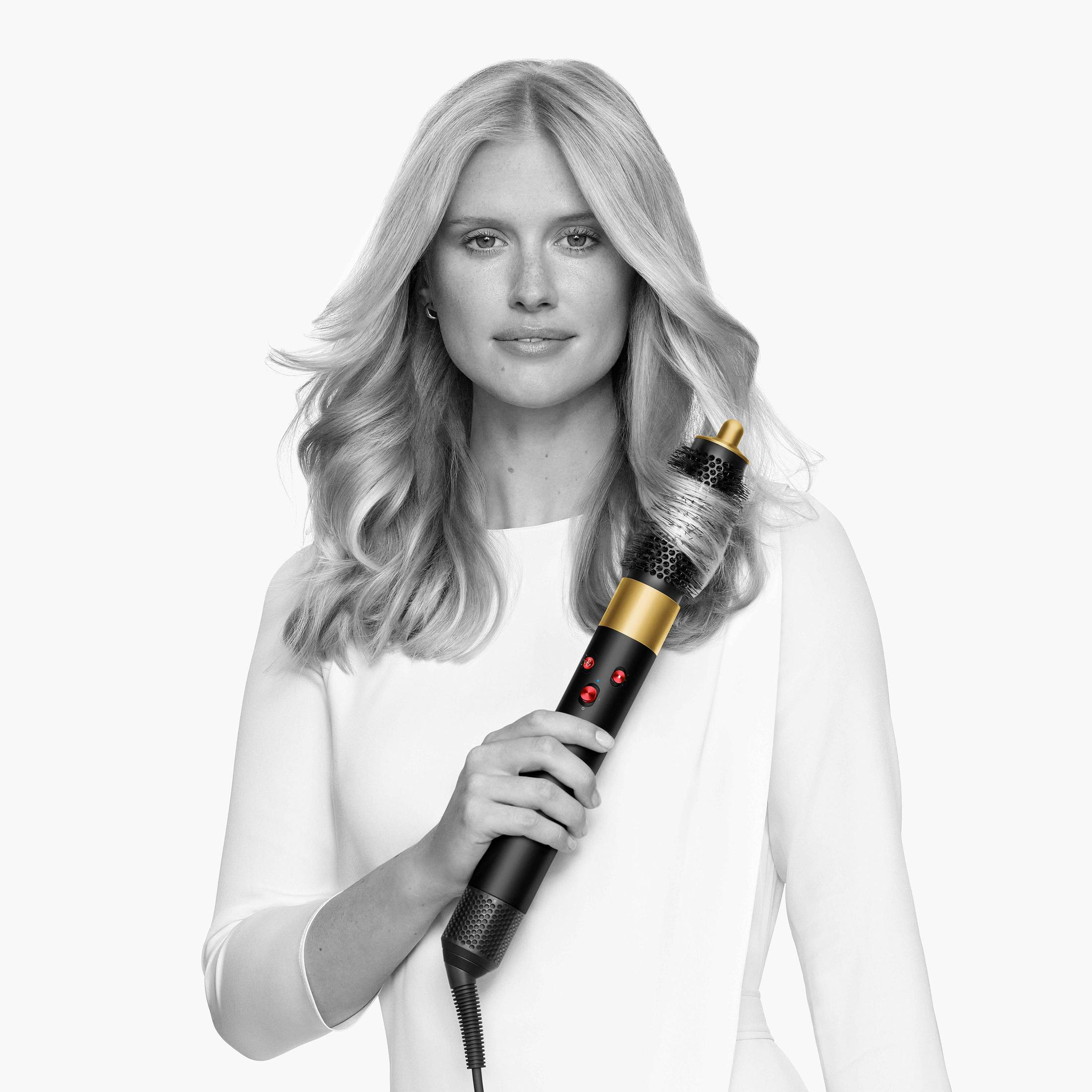 Frau hält eine Haarstyling-Bürste, heller Hintergrund. Die Bürste ist schwarz und gold, mit roten Knöpfen.