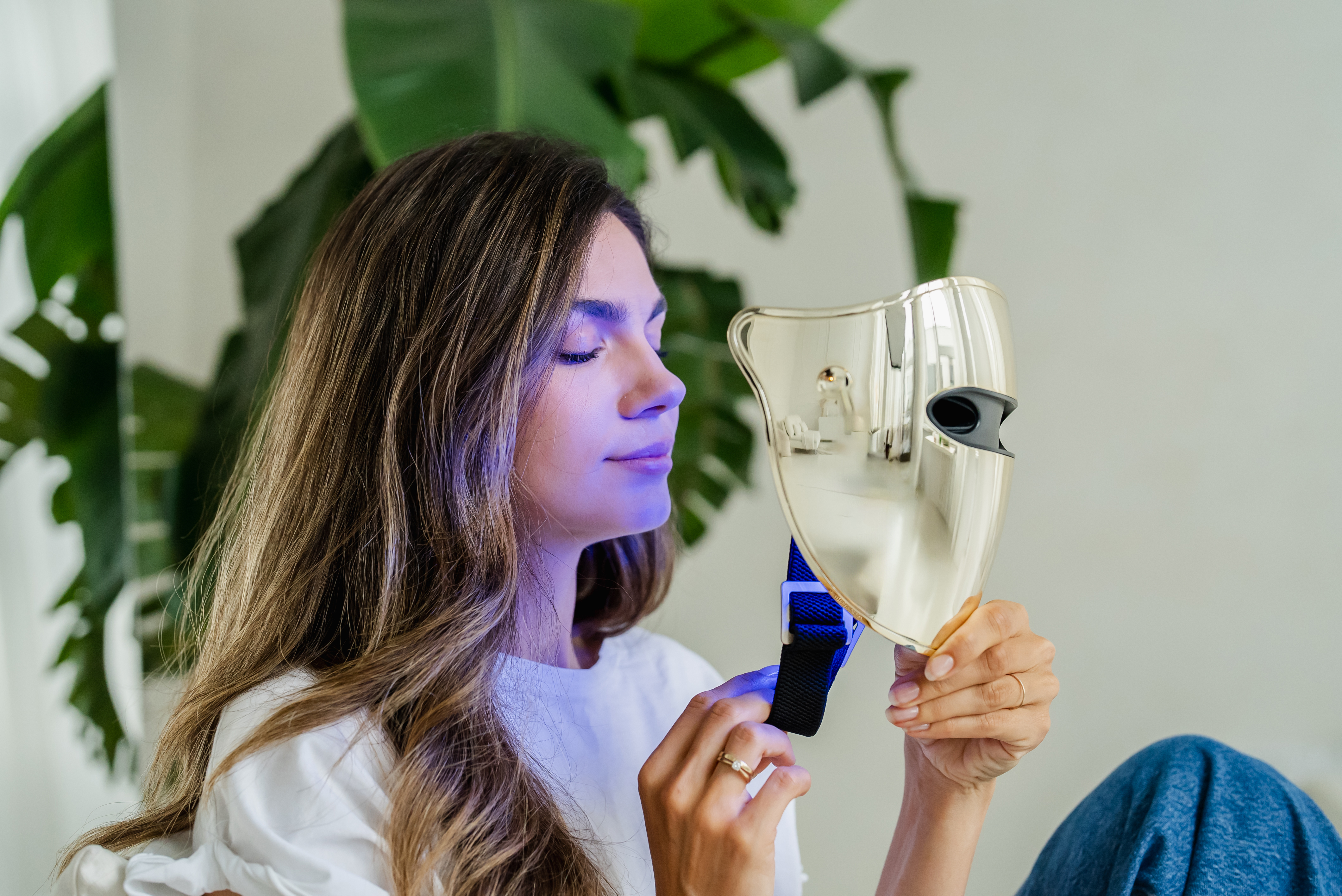 Una donna tiene una maschera facciale dorata, con luce blu, davanti a piante verdi.