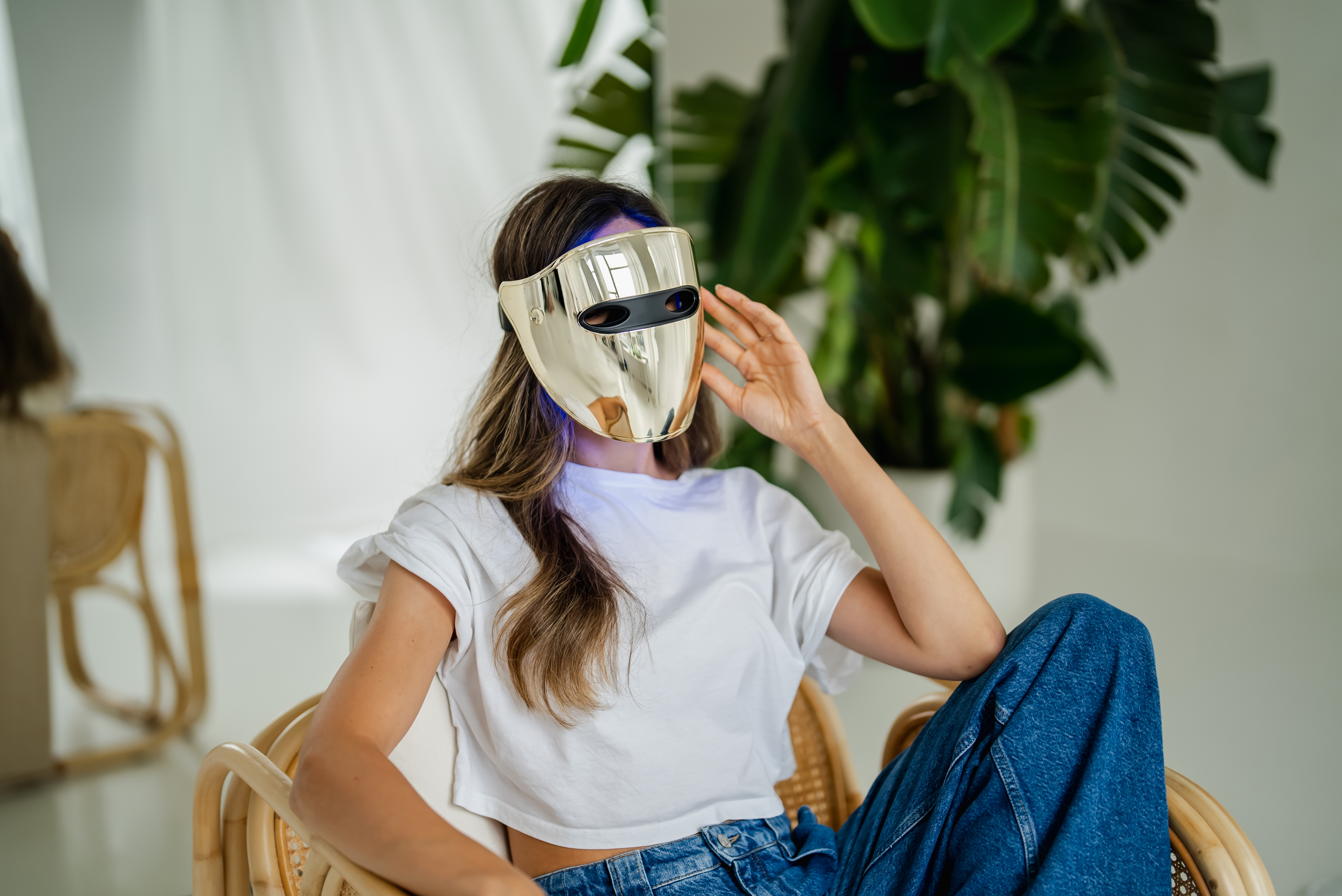 Una donna con una maschera facciale LED dorata, seduta su una sedia, con una pianta verde sullo sfondo.