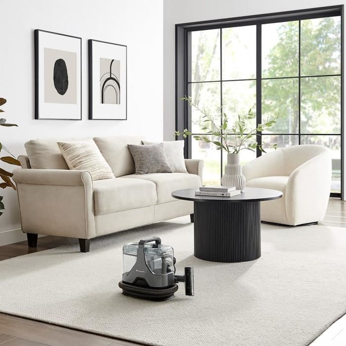 Wohnzimmer mit hellem Sofa, weißem Stuhl, rundem schwarzen Tisch und einem Staubsauger.