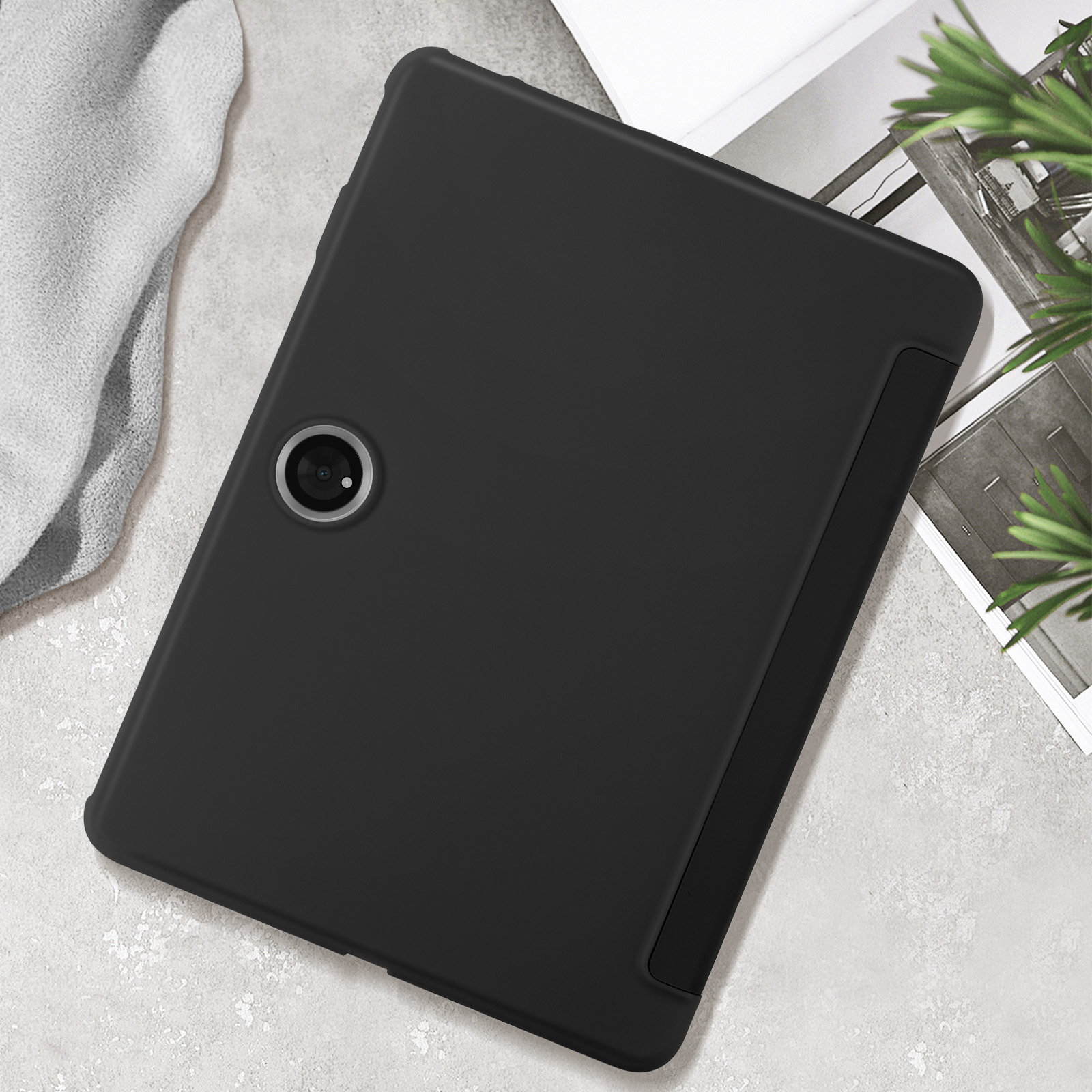 Custodia per tablet nera con foro per fotocamera. Si trova su una superficie grigio chiaro con un foglio bianco e foglie.