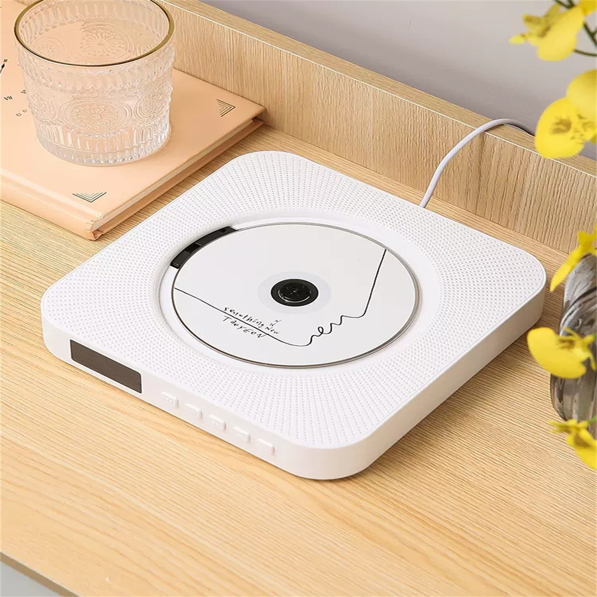 Weißer CD-Player, ein Buch und ein Glas auf Holz. Gelbe Blumen im Hintergrund.