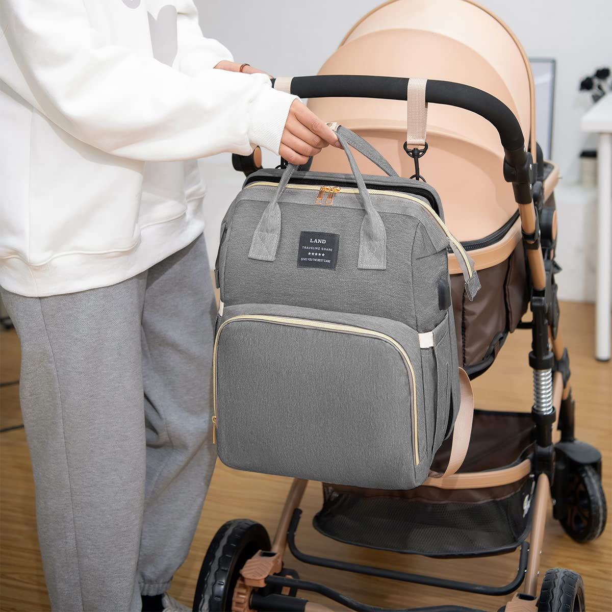 Person hängt eine graue Wickeltasche an einen Kinderwagen. Die Tasche hat einen Griff und einen Riemen.