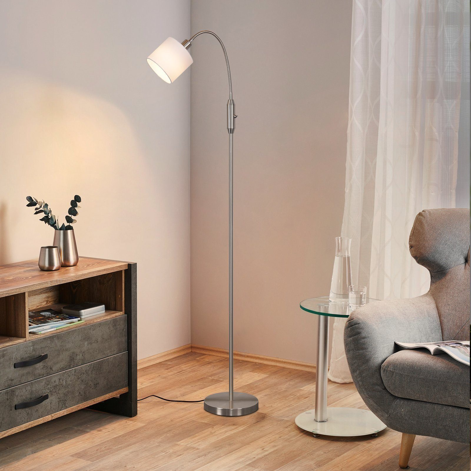 Eine hohe, silberne Stehlampe mit weißem Lampenschirm steht in einem Raum mit Holzschrank und grauem Sessel.