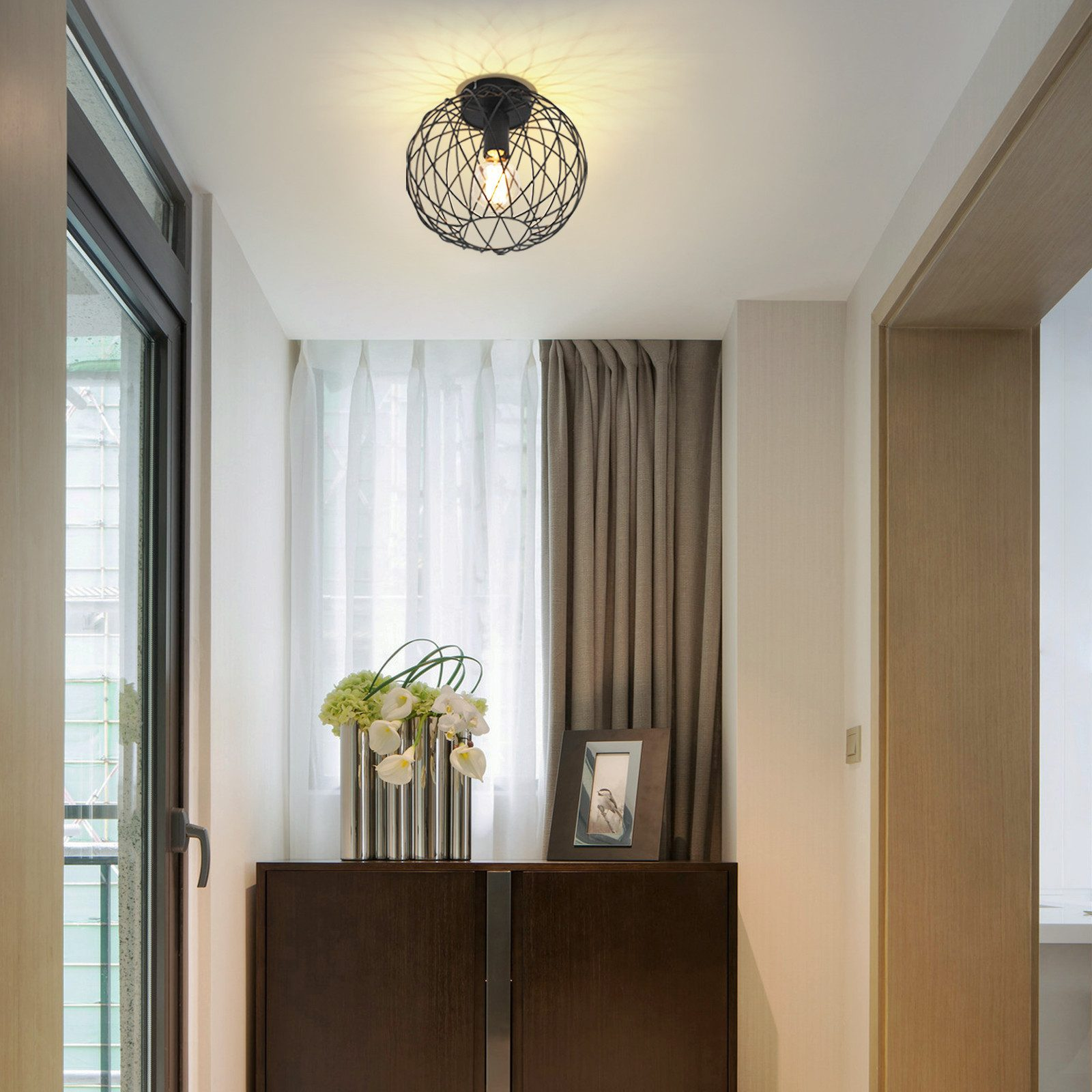 Un corridoio con un lampadario. Un mobile con un vaso e una cornice. Tende e una finestra sono visibili.