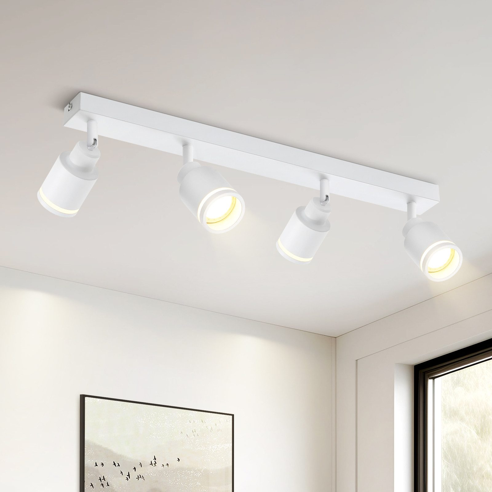 Apparecchio di illuminazione a binario bianco con quattro faretti orientabili. Montato su un soffitto bianco. Una parete sullo sfondo.